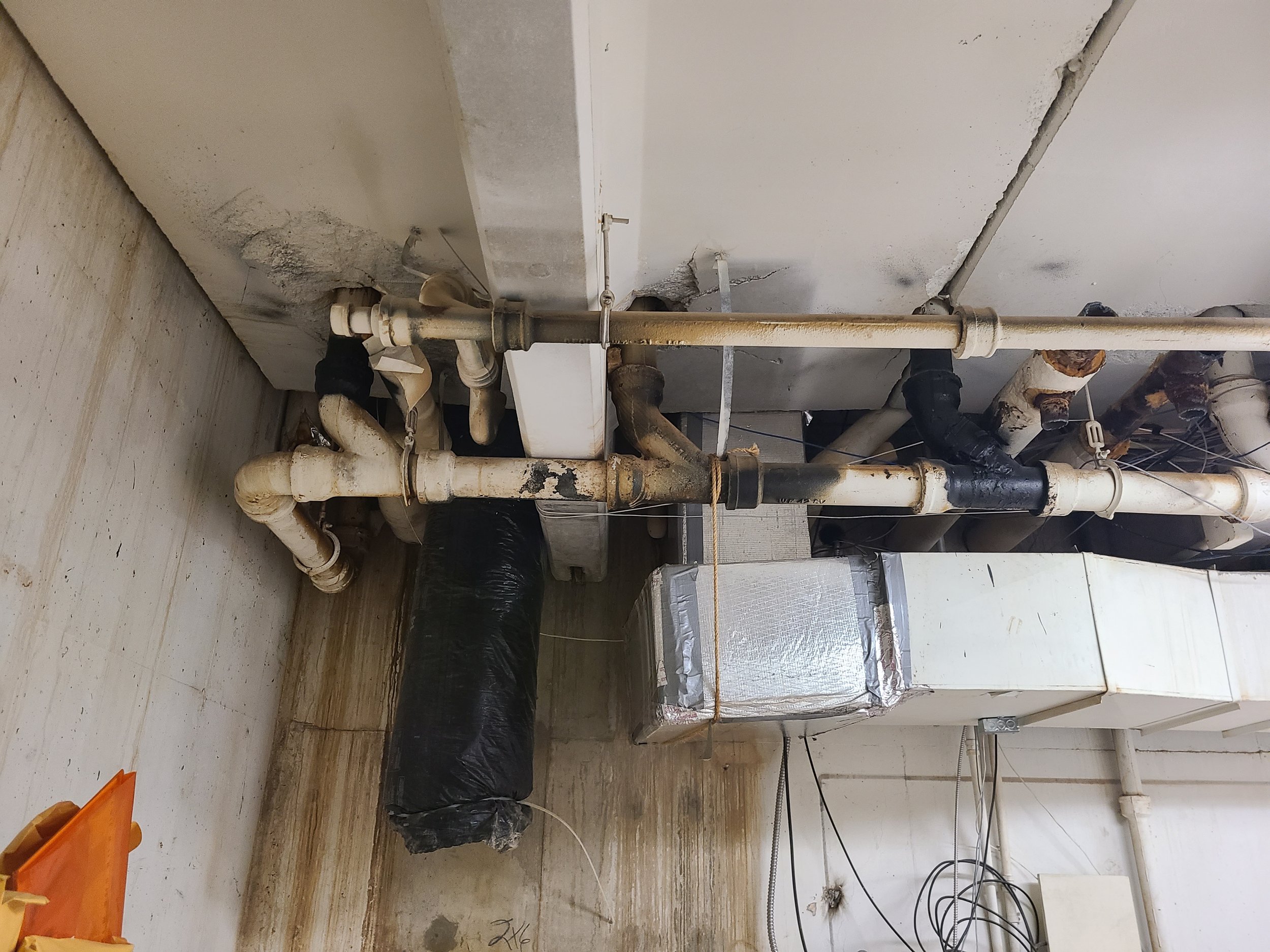 Pipes and plumbing in a wall under construction or repair, with some debris and black plastic covering on the floor.