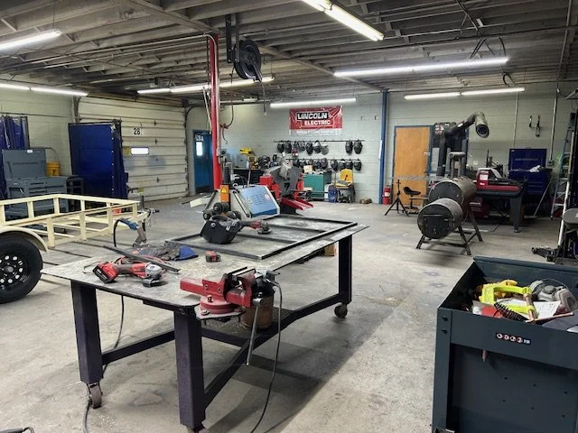 An industrial workshop with various tools and equipment, including a worktable with a grinder, a bandsaw, and safety gear, illuminated by ceiling lights.