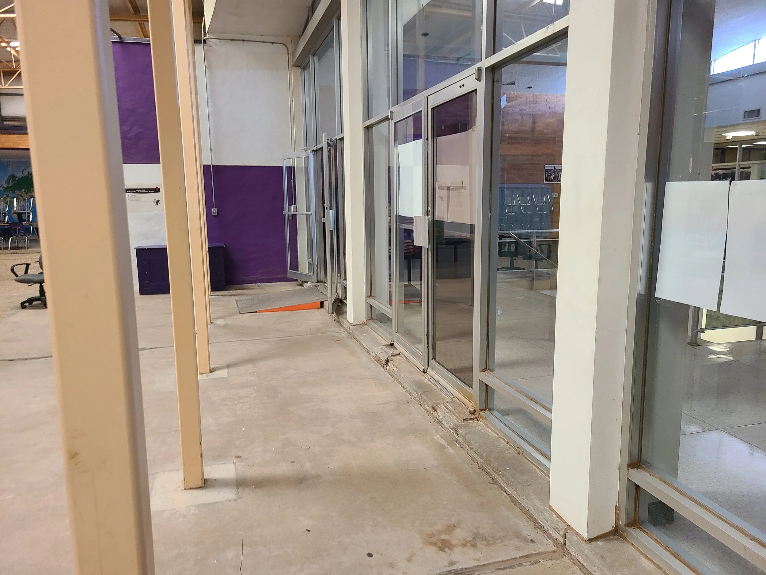 Interior of a building under construction or renovation, with exposed concrete floor and framing for glass walls or doors, a purple wall section, and some chairs stacked in the background.