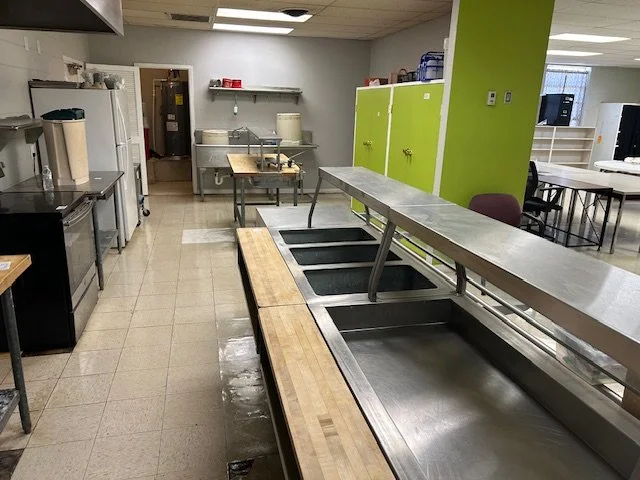 Empty cafeteria or kitchen serving area with stainless steel counters, lockers, and some kitchen equipment in the background.