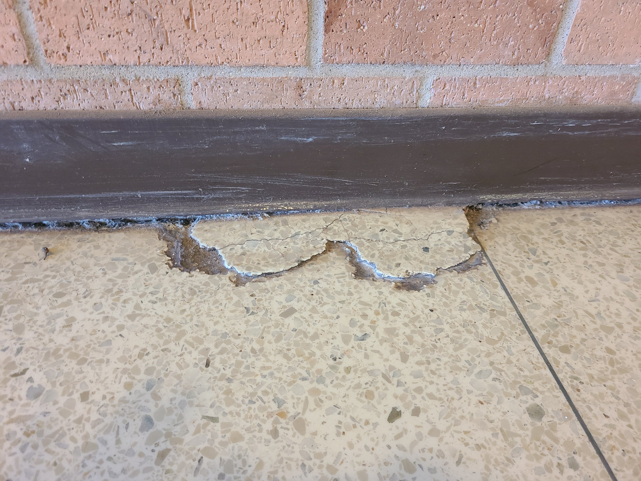 Close-up of a brick wall with a black baseboard at the bottom, next to a speckled tile floor with a crack.