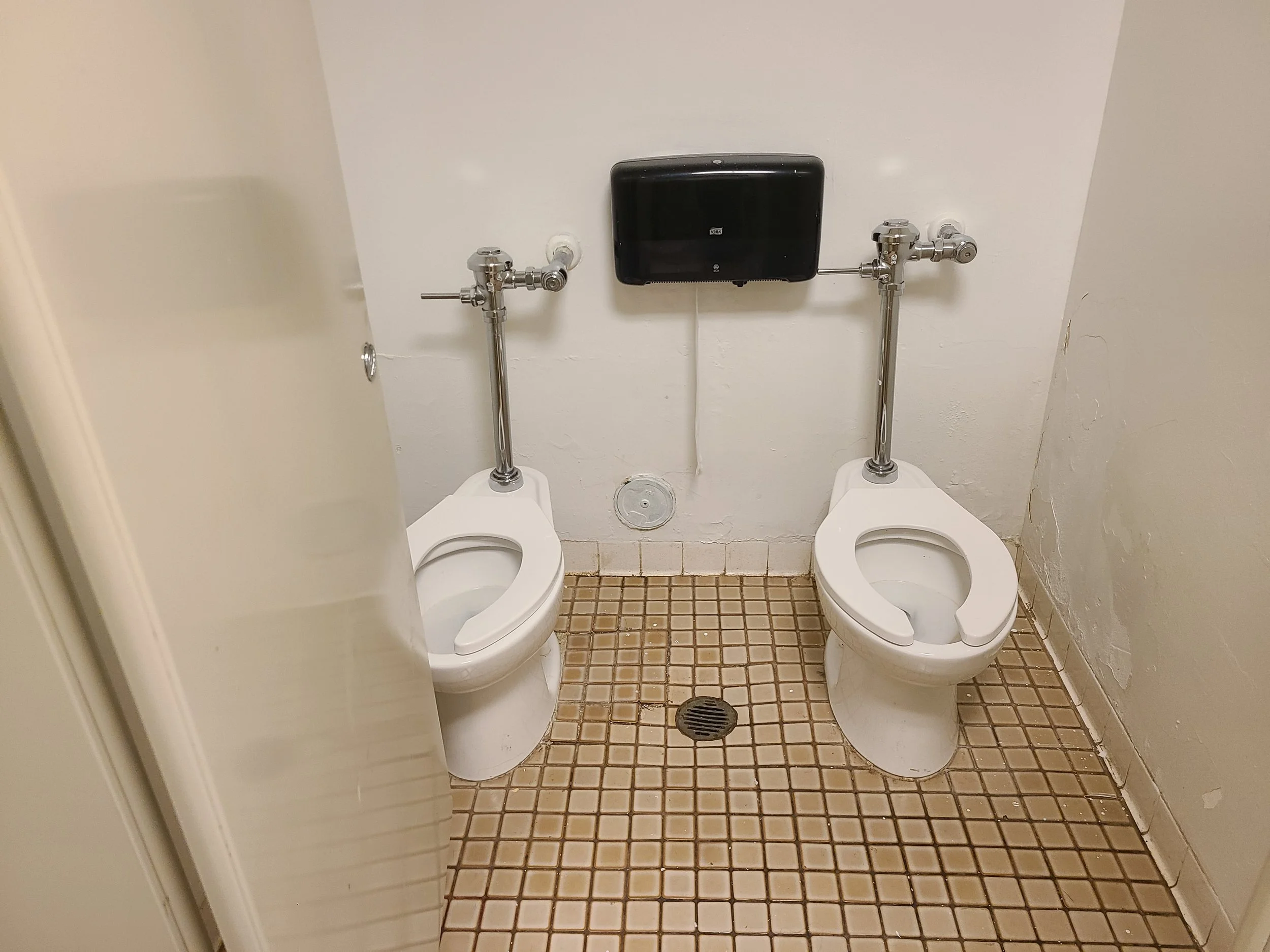Public restroom with two white toilets side by side, metal flush pipes, a black box on the wall, a floor drain, and beige tiled floor.