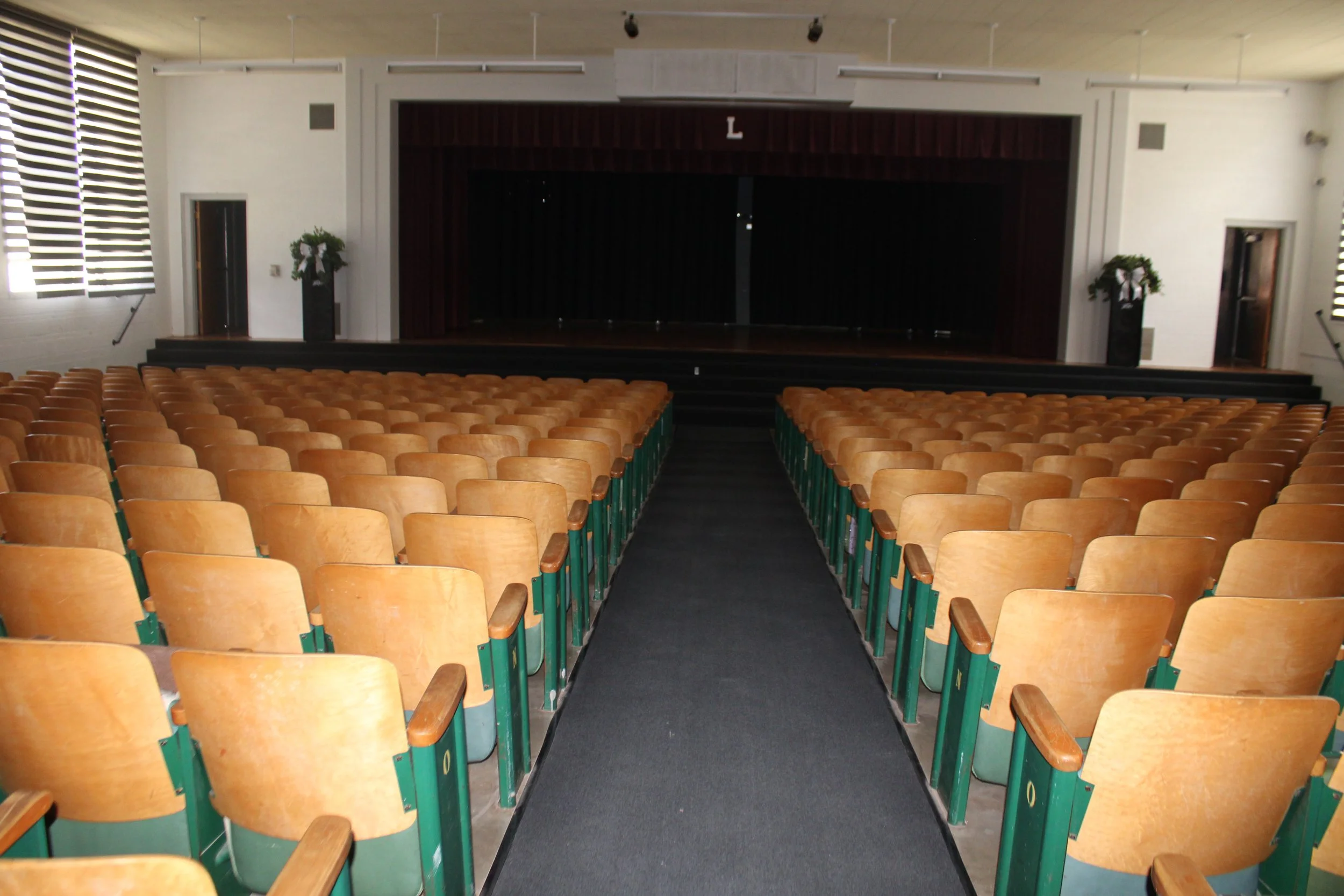 An empty auditorium with rows of wooden chairs facing a stage with dark curtains, decorated for a holiday with two wreaths on tall black vases, and large windows with horizontal blinds on the side walls.
