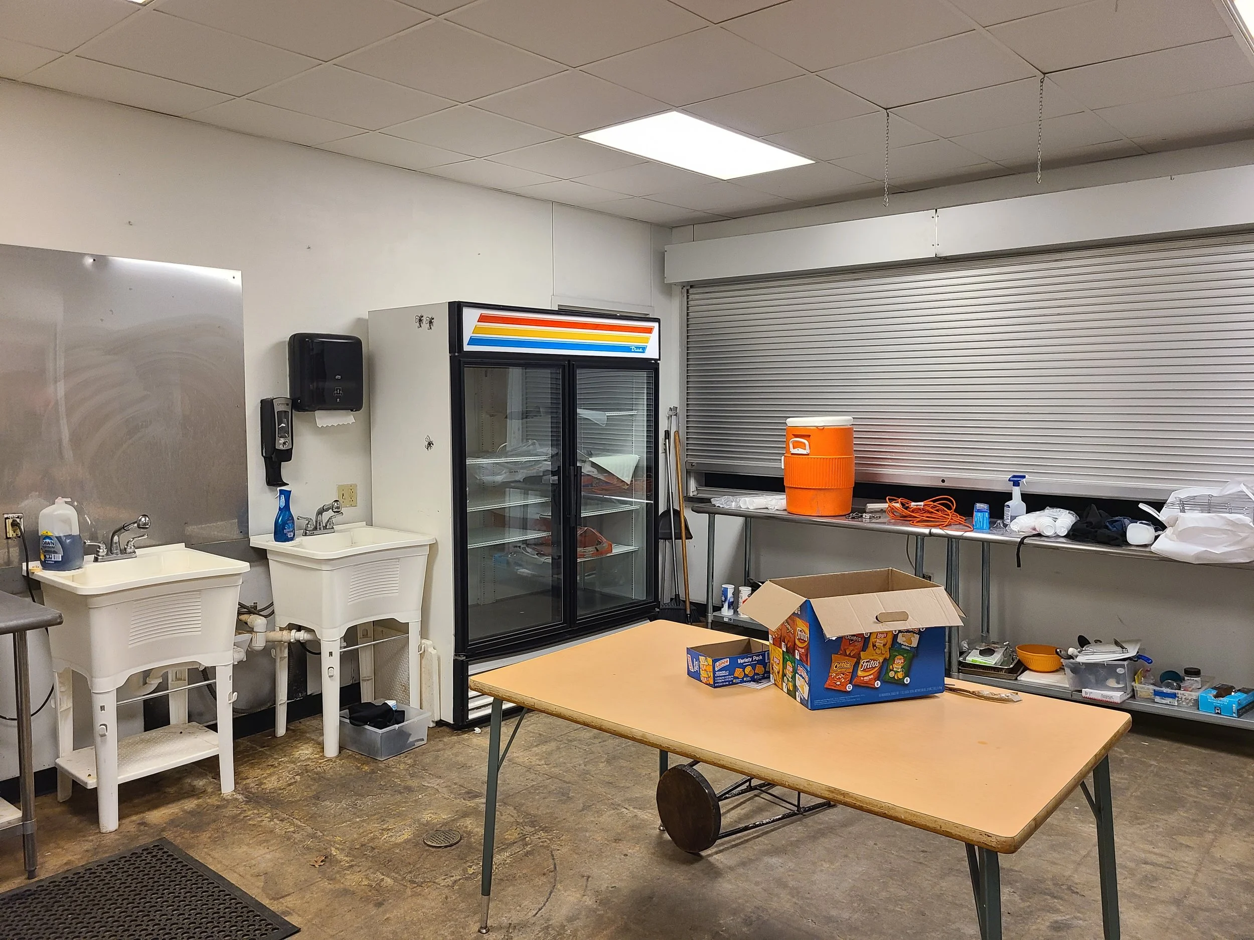 A cluttered room with white walls and a drop ceiling. There are two sinks with soap and cleaning supplies, a black paper towel dispenser, a glass-front refrigerator with a colorful striped logo, an orange water cooler, a table with snack boxes, a car