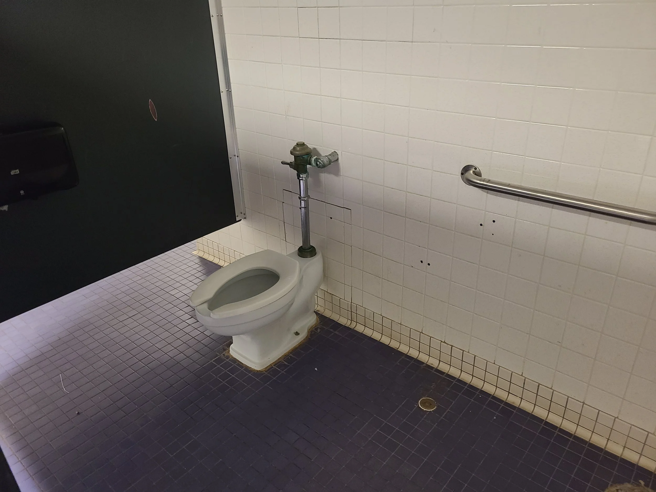 A public restroom with a wall-mounted toilet and a water pipe, black partition, and blue tiled floor.