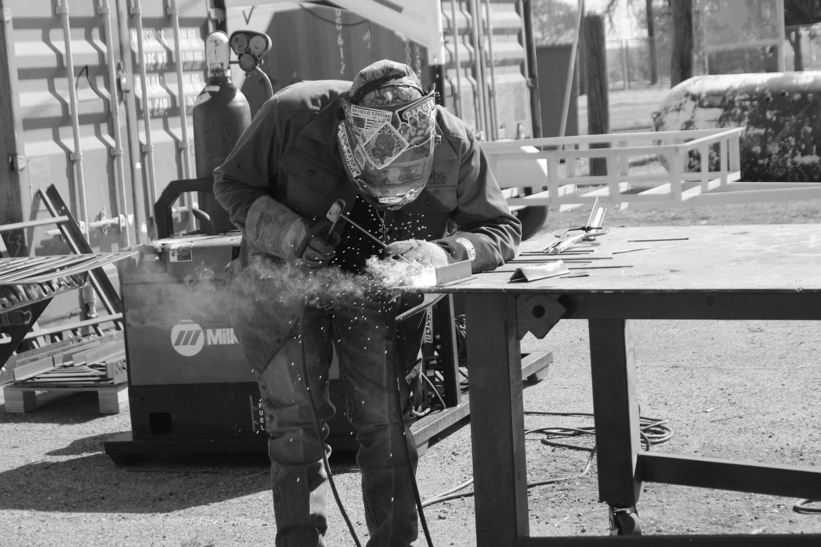 A welder welding metal at an outdoor workshop, with sparks flying, wearing protective gear including a helmet and gloves.
