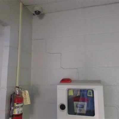 A fire extinguisher mounted on a concrete wall next to a cabinet with a glass door, with a red object on top, in a room with a security camera in the corner.