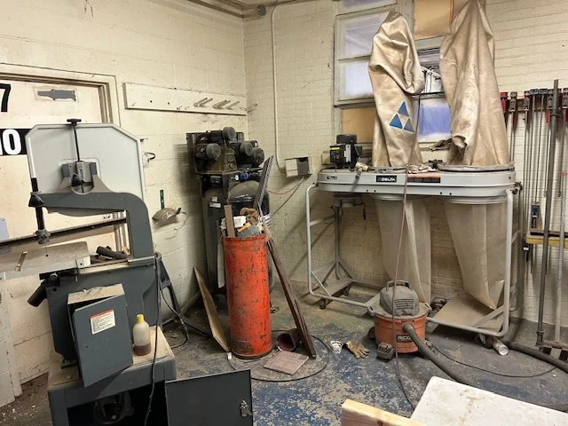 Workshop with various tools, equipment, and machinery, including a bandsaw, an orange gas cylinder, a worktable covered with cloth, and power tools, in a cluttered space.