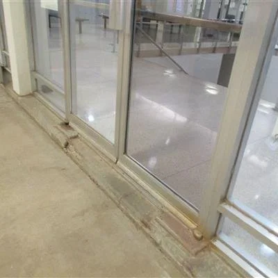 Close-up of a dirty, sealed glass door with a worn baseboard and a concrete floor, showing signs of dirt and damage at the bottom of the frame.