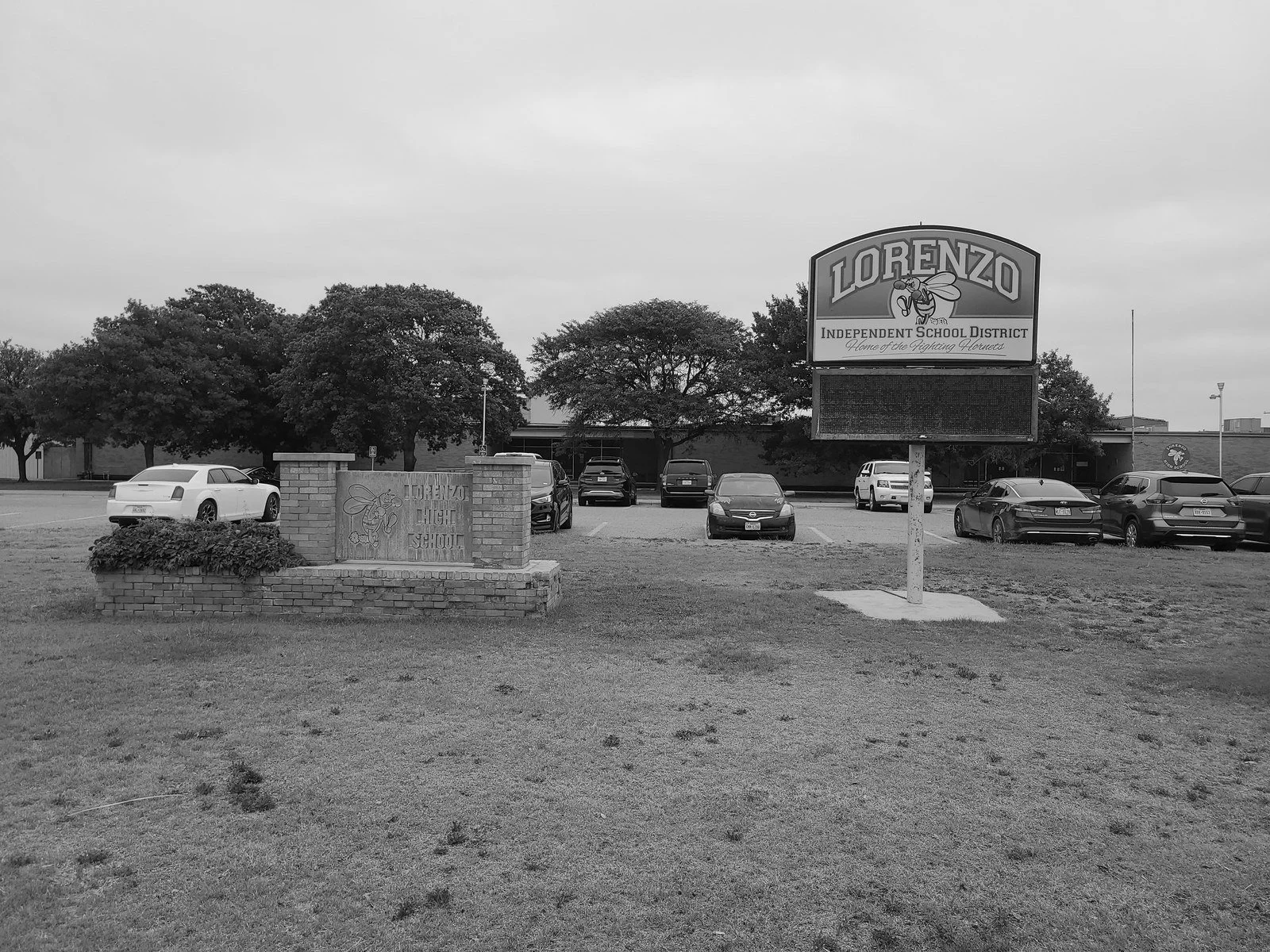 Black and white photo of the Lorenzo High School sign and parking lot with several cars and trees in the background.