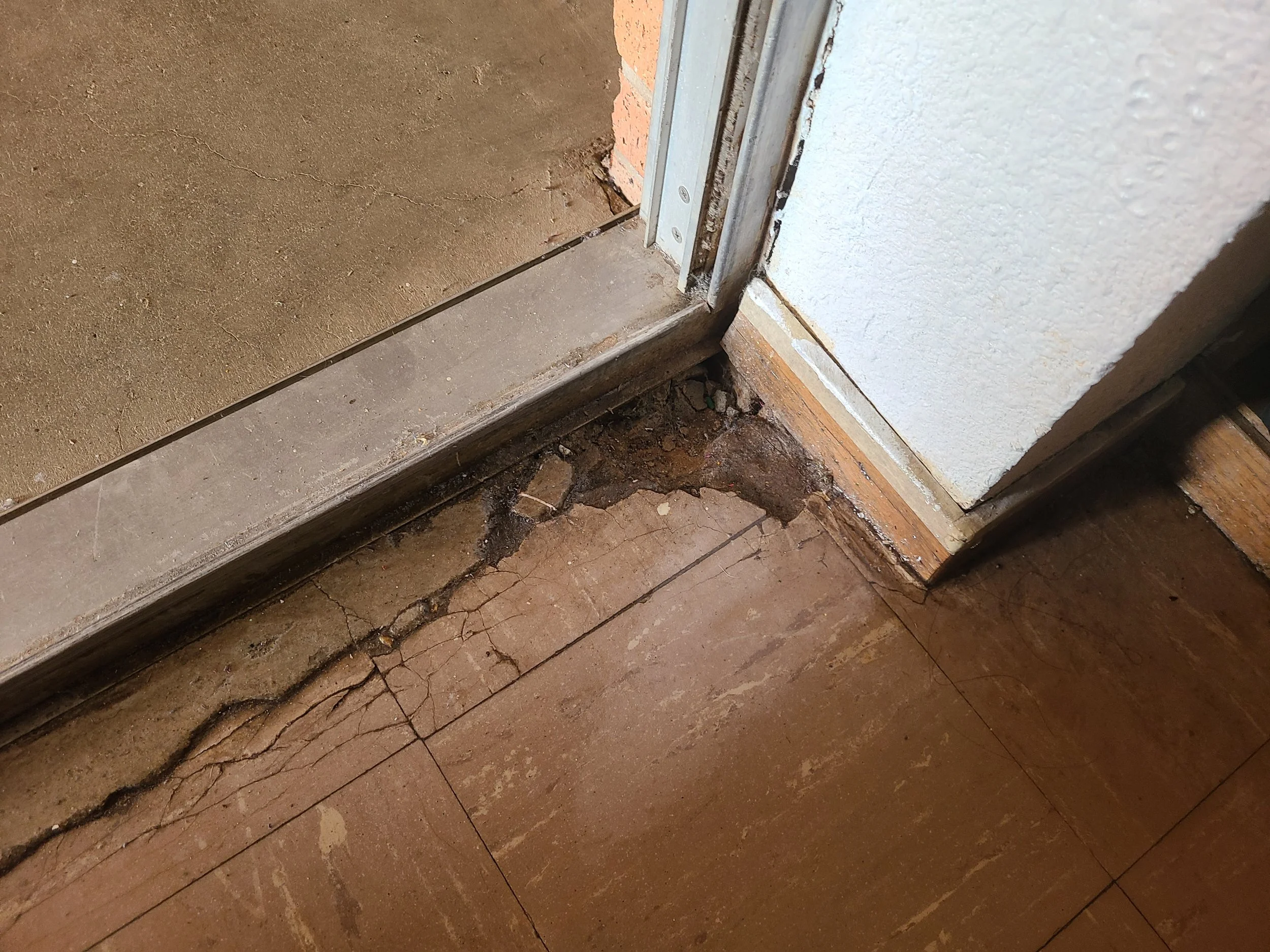 Close-up of a wooden floor, showing cracked and damaged wood near a door threshold with metal framing, revealing the subfloor underneath.