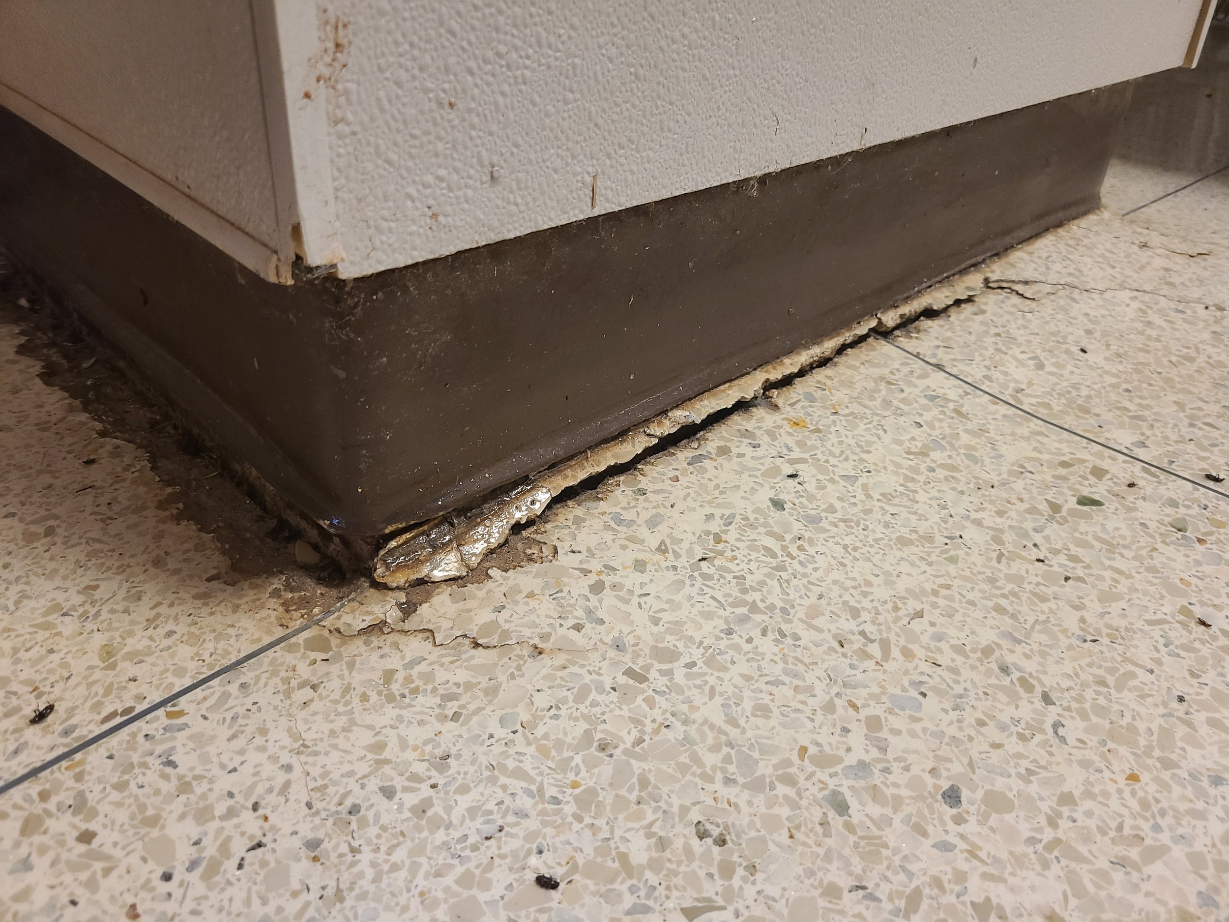 Close-up of a damaged kitchen or appliance base with chipped and broken edges, showing a layer of what appears to be melted or damaged material, on a terrazzo floor.