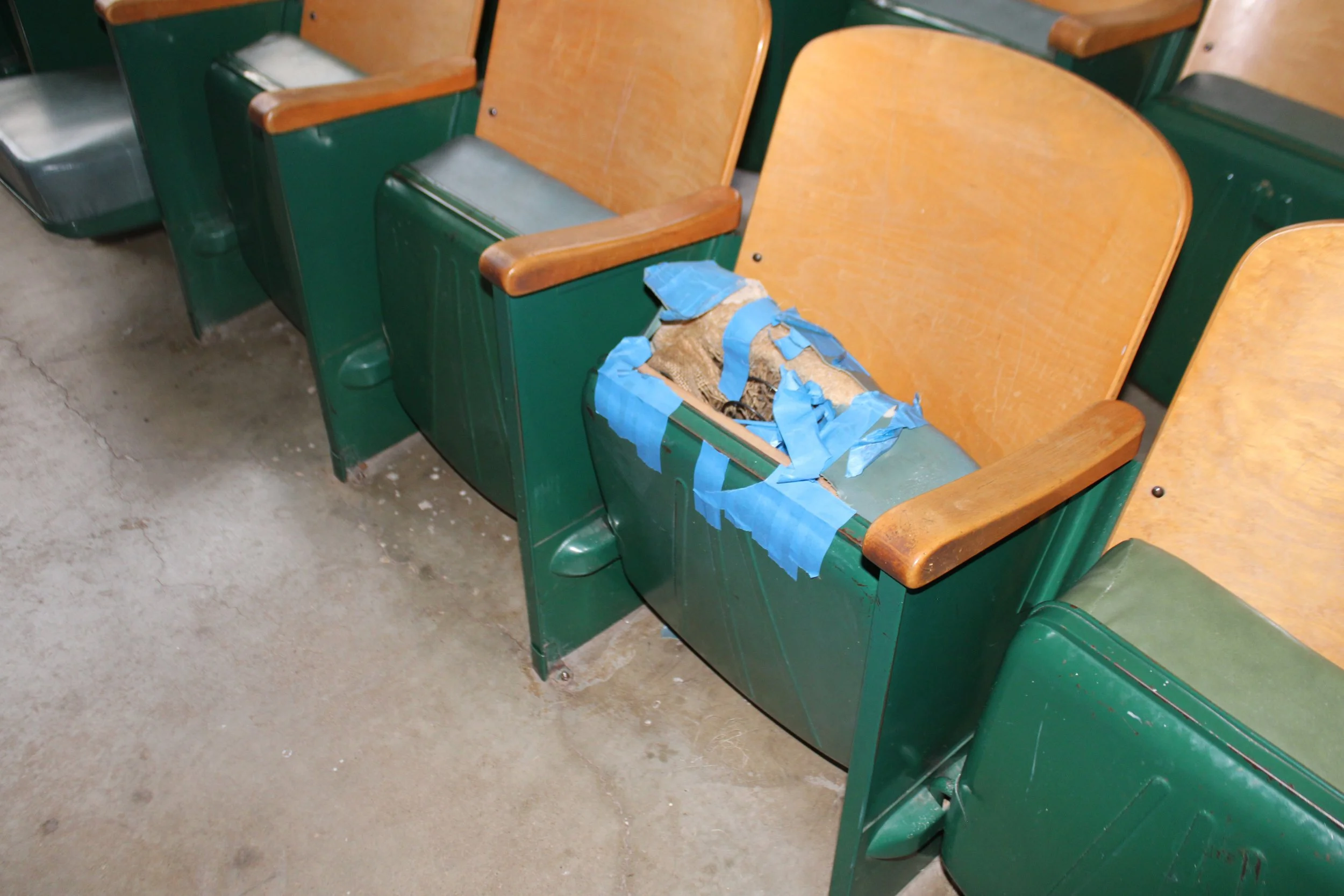 Empty green theater seats with wooden armrests, damaged with torn blue tape and a hole revealing stuffing inside.