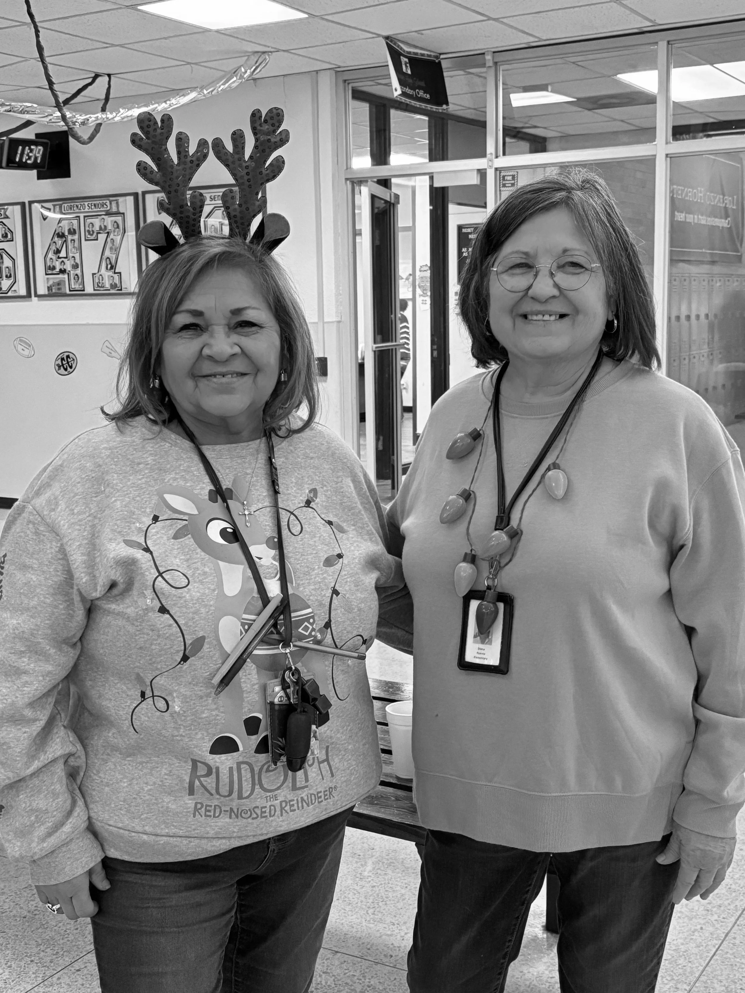 Two women standing together indoors, one wearing reindeer antlers and a Rudolph-themed sweatshirt, and the other wearing glasses and a necklace. Both are smiling and wearing lanyards with ID badges.