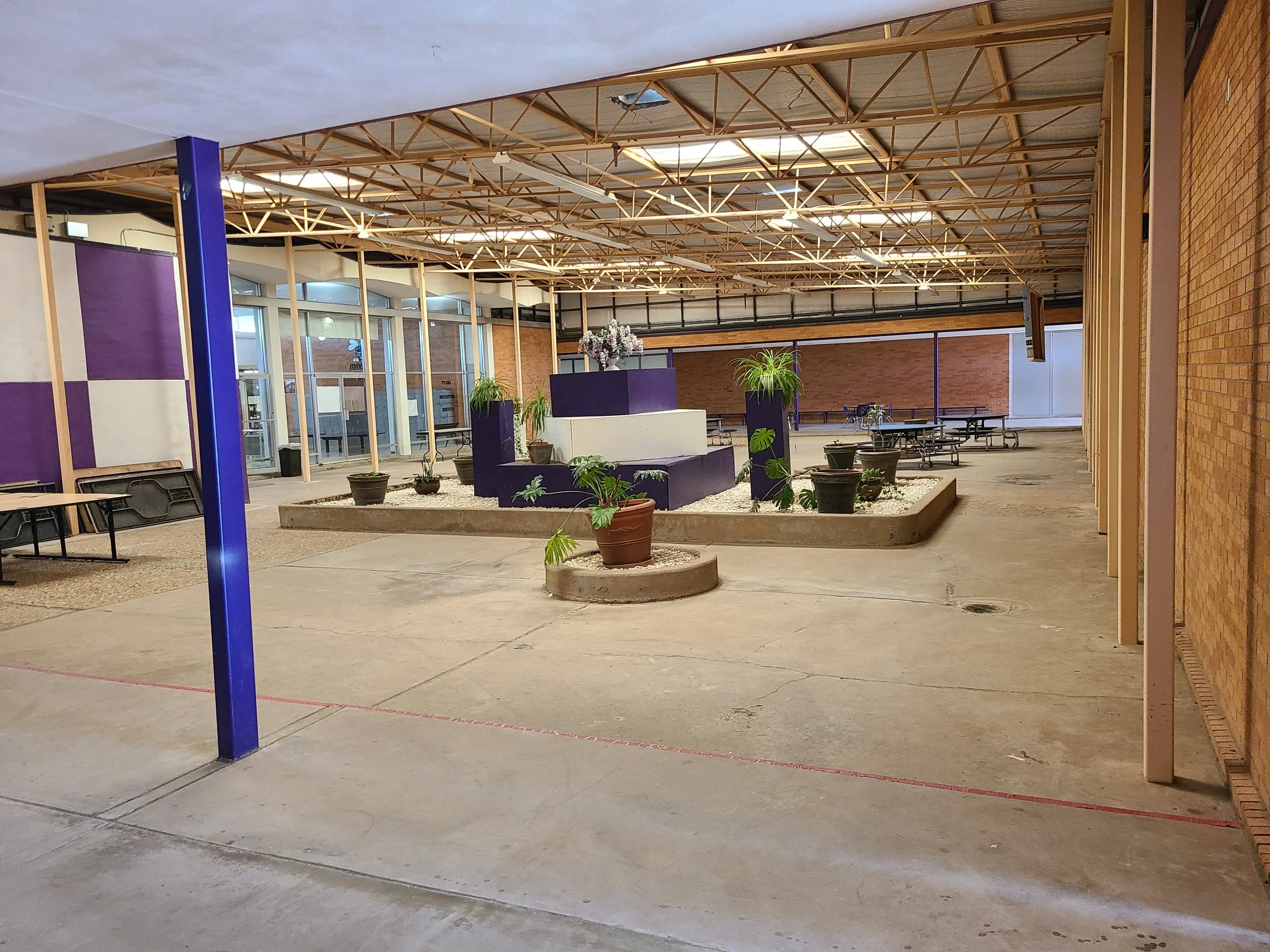 Interior of a building with a high ceiling under construction, showing wooden framework, potted plants, benches, and a brick wall on the right side.
