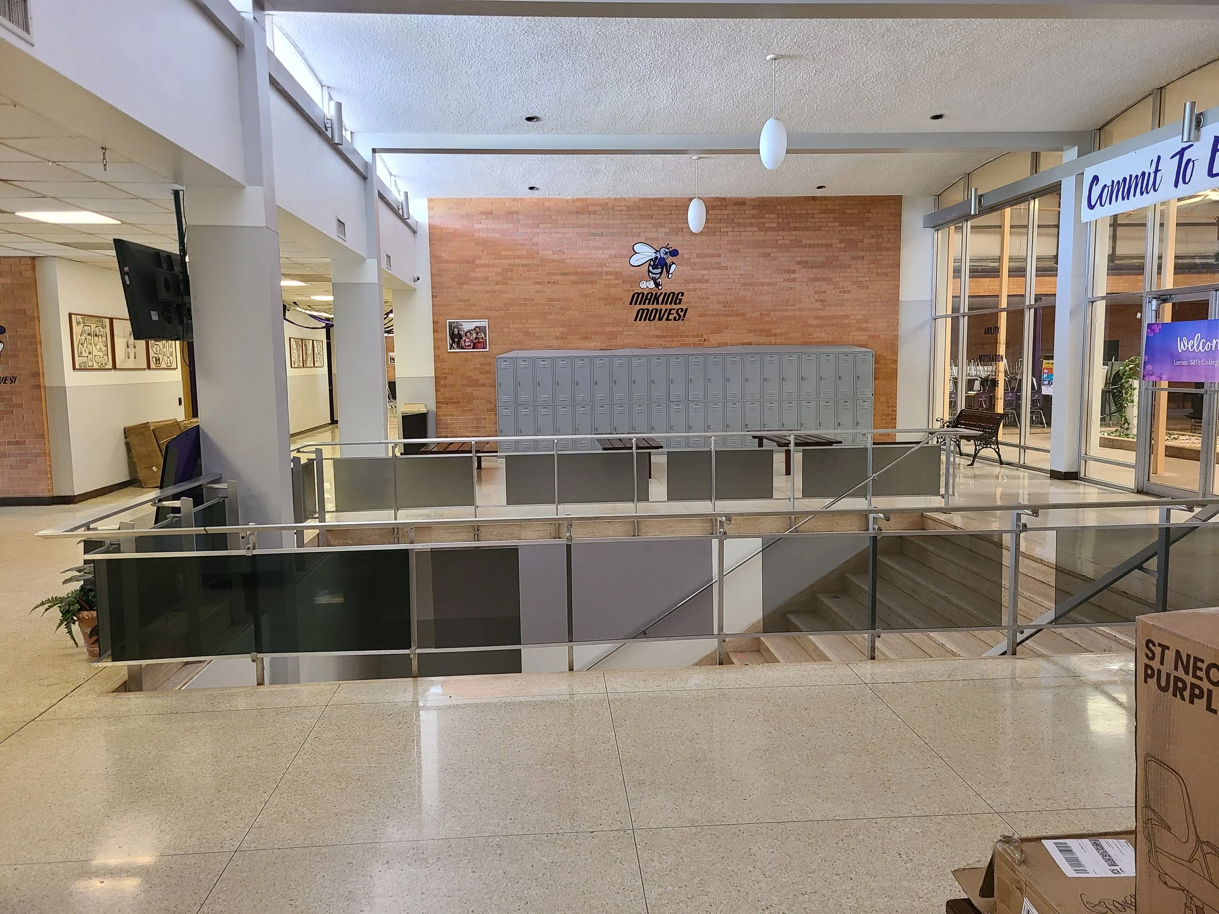Interior view of a school or college corridor with lockers on a brick wall, a motivational bee logo, and a sign that reads 'Commit to...', with seating areas, a staircase, and various posters and signs.