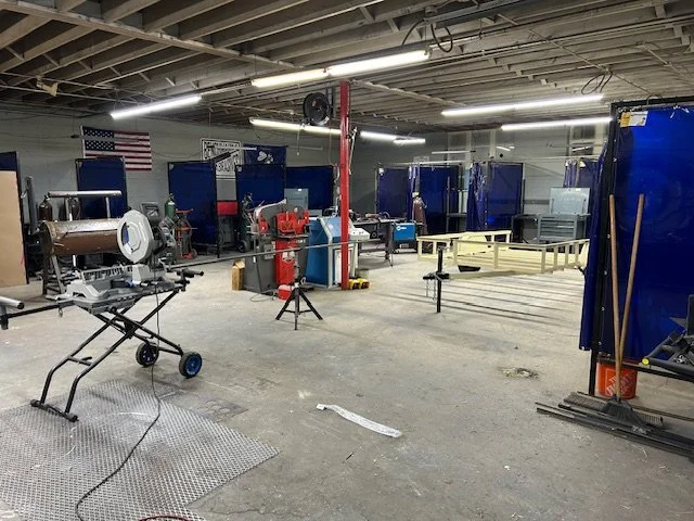 A large industrial workshop with welding stations separated by blue curtains, welding equipment, and tools, with tools on wheeled carts and American flag on the wall.