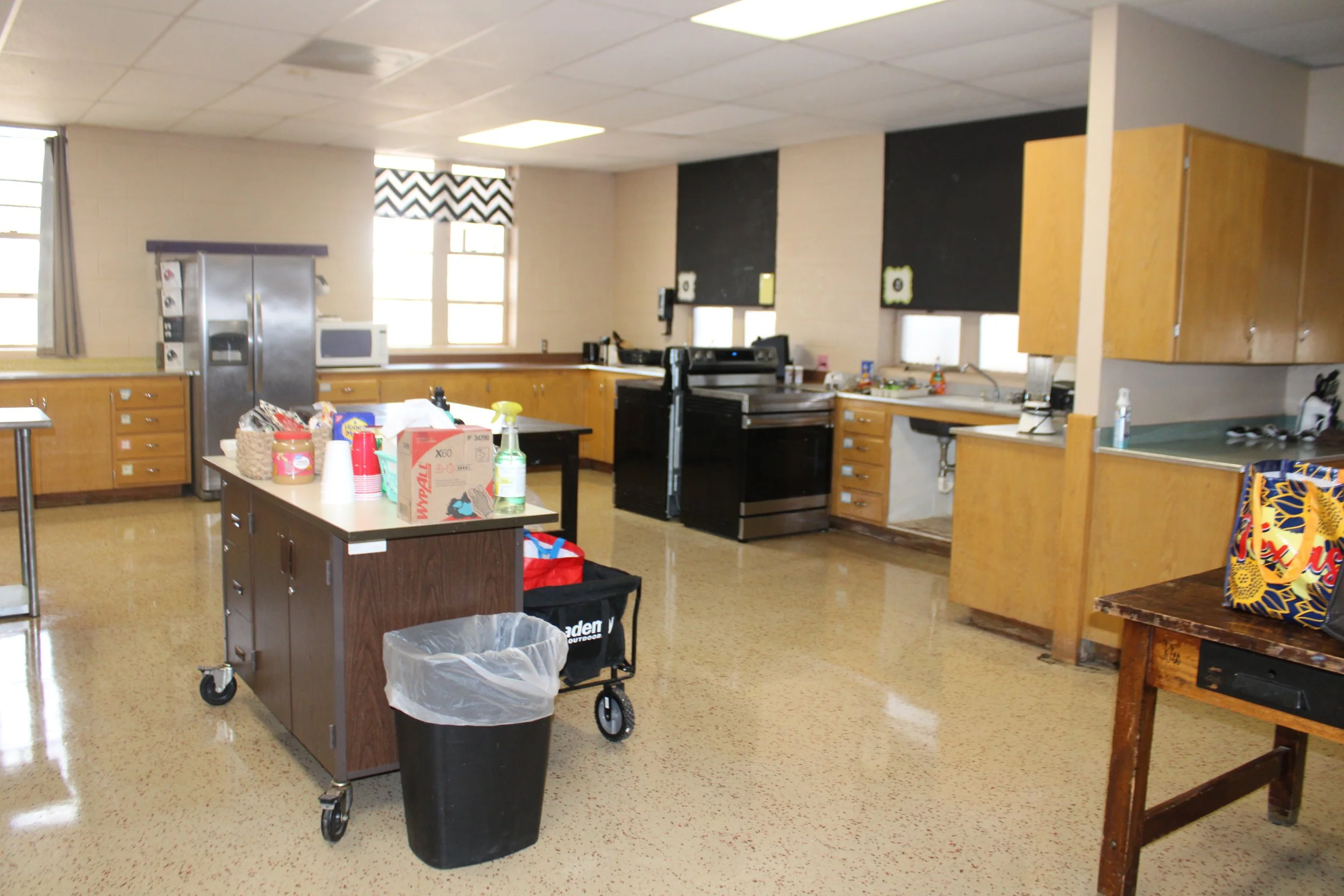 A kitchen or common room with wooden cabinets, a black stove, microwave, and large windows with black and white zigzag curtains. There are various items on the counters and a rolling cart with supplies and trash.