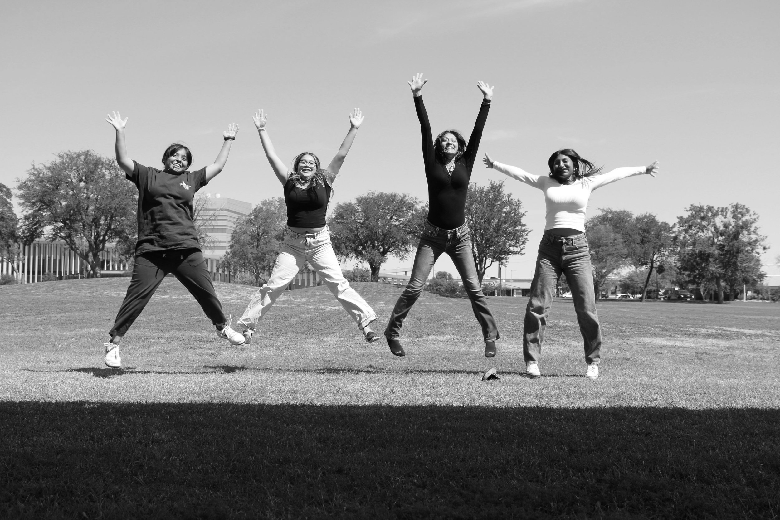 Four women jumping in the air on a grassy field in a park with trees and buildings in the background.