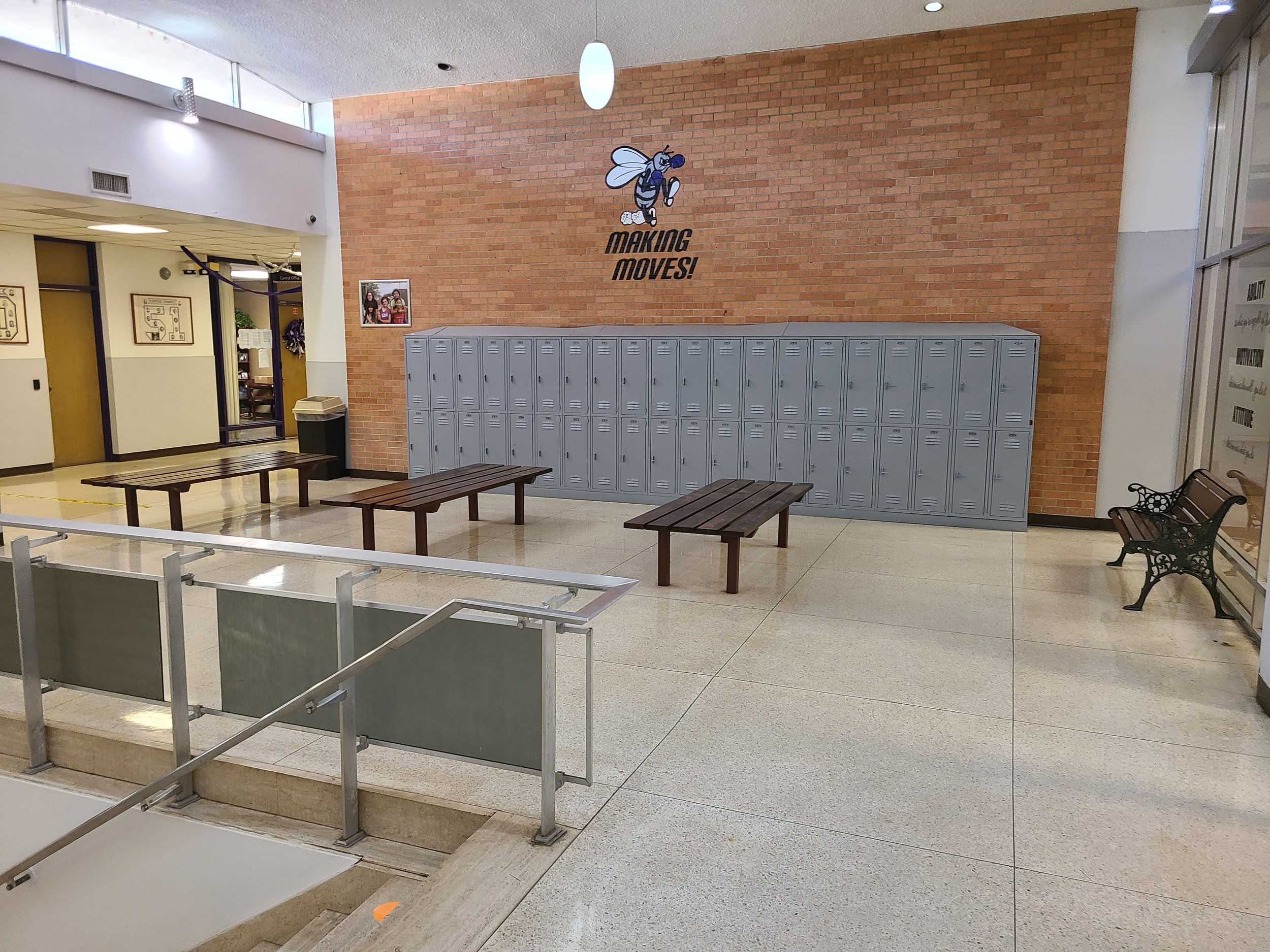 Interior of a school hallway with gray lockers, benches, and a brick wall with a bee mascot and the text "Making Moves!"