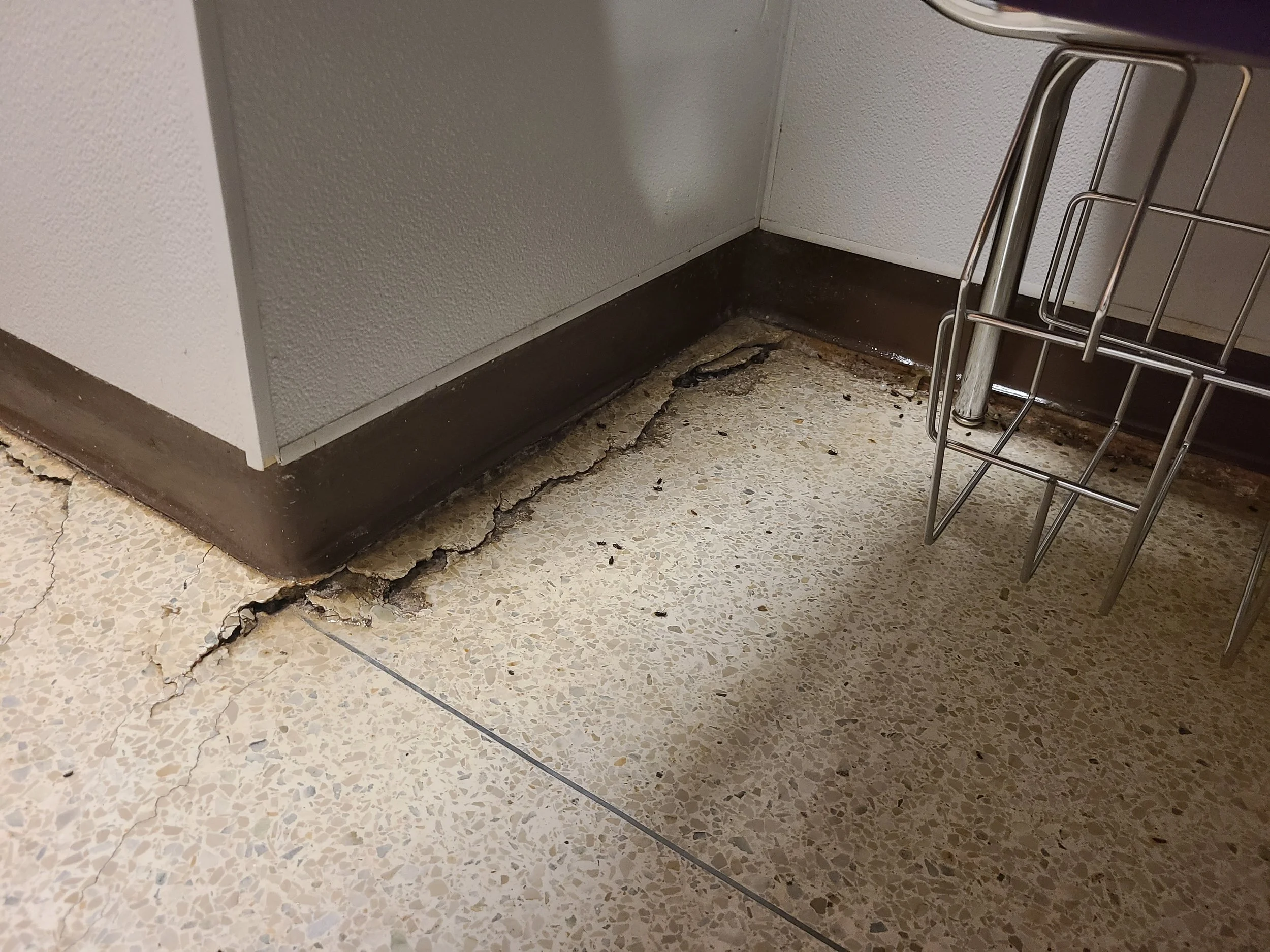 Floor with cracked tile and a damaged baseboard corner, next to a white wall and a metal rack.