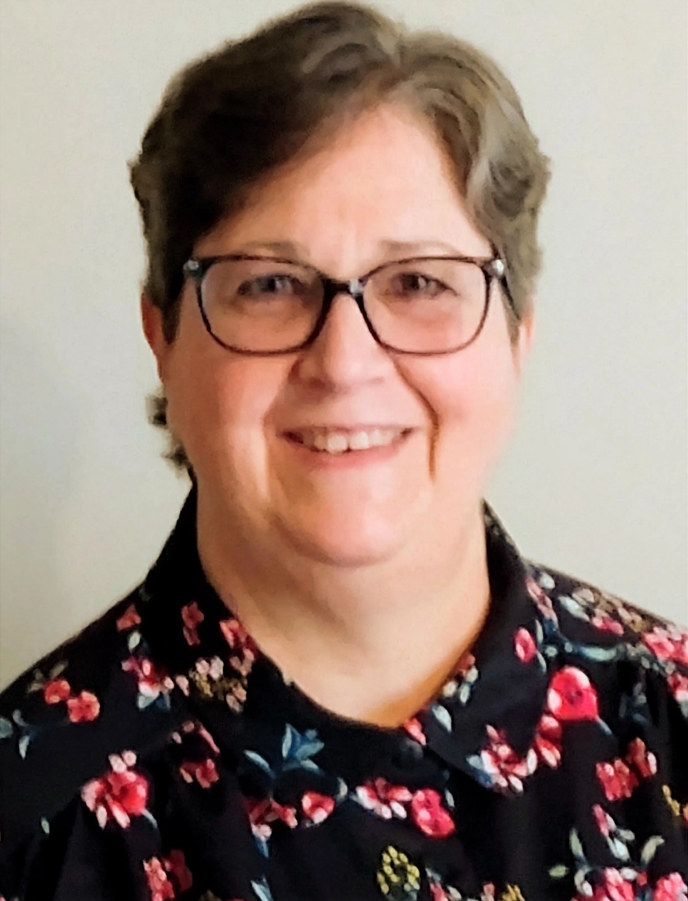 Close-up of a smiling woman with short, brown hair, glasses, and wearing a black top with a vibrant floral pattern.
