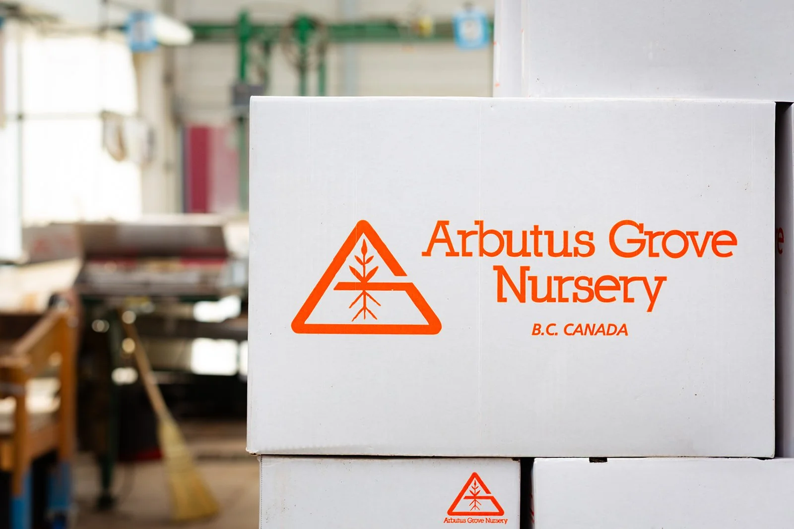 White boxes with orange logo and text reading 'Arbutus Grove Nursery B.C. Canada' in a nursery or warehouse setting.