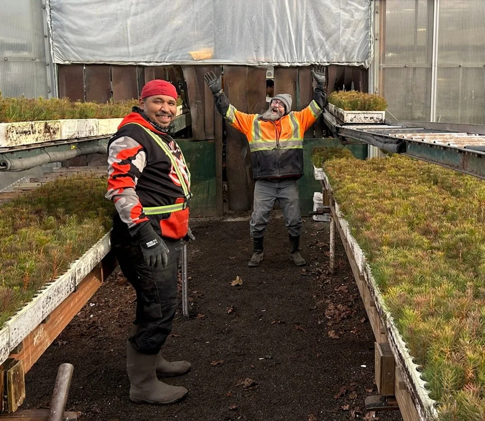 Two men inside a greenhouse, surrounded by young plants on shelves. The man on the left wears a red hat and a black and red jacket, smiling, with rubber boots and gloves. The man on the right has his arms raised in celebration, wears glasses, a beani