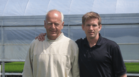 Hans and Nathaniel  standing outdoors near a greenhouse, with one older man wearing a light sweater and a younger man in a black shirt, both smiling.