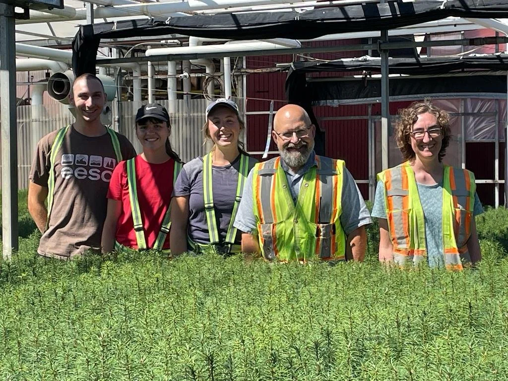 Group of five people standing outdoors behind a row of green plants, smiling, with industrial structures in the background. Four are wearing safety vests, and one person is wearing a hat and sunglasses.