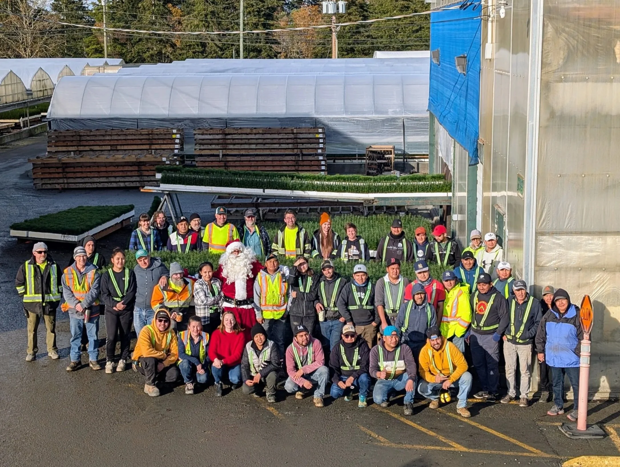 A group of people standing in front of greenhouses, some dressed in safety vests, with a person dressed as Santa Claus among them. The setting appears to be a farm or plant nursery.