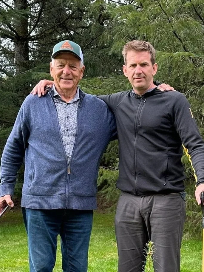 Hans and Nathaniel standing outdoors in front of trees, with their arms around each other's shoulders, smiling at the camera.