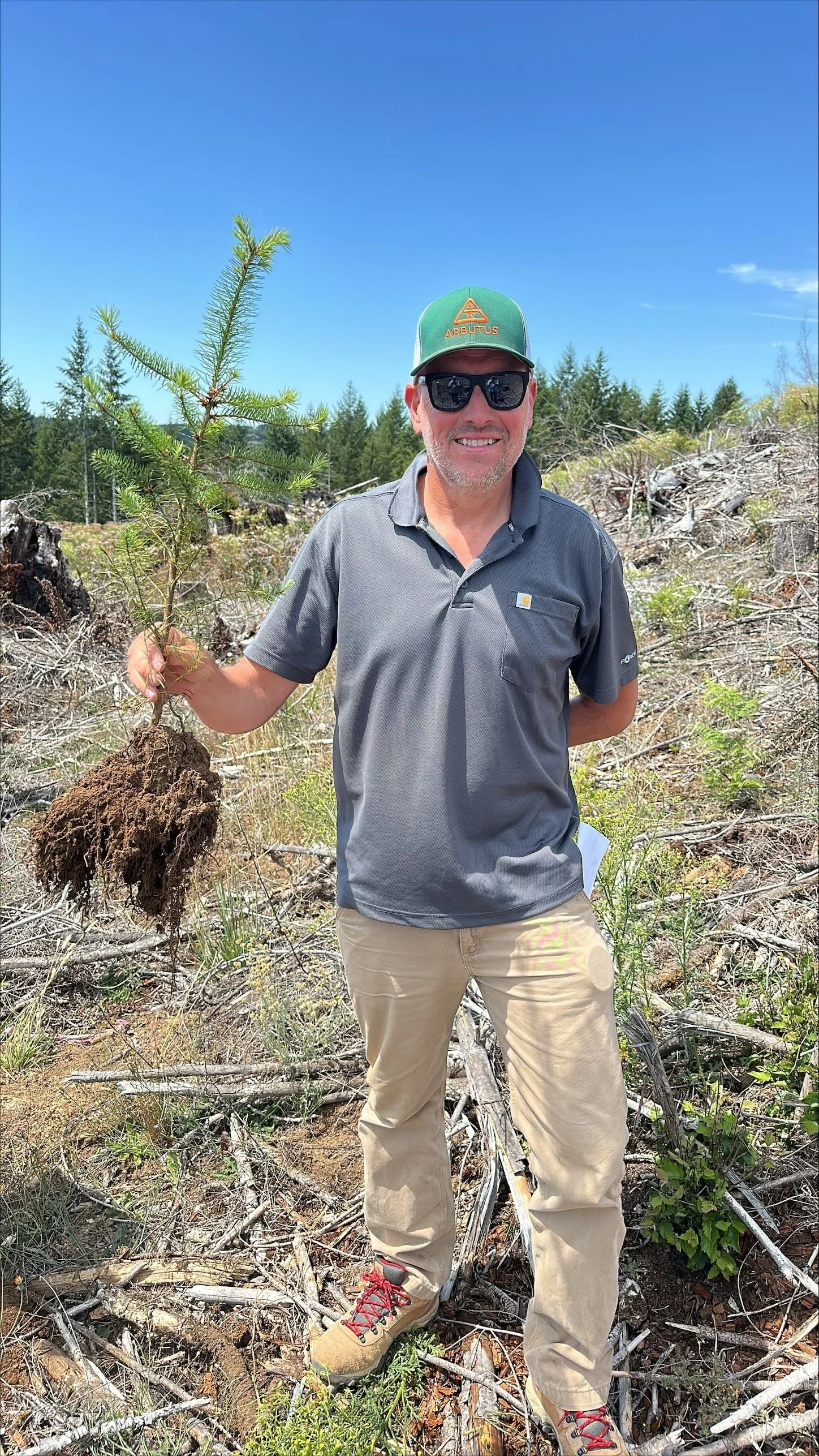 A man in outdoors in a forested area holding a small uprooted pine tree with its roots exposed. He is wearing sunglasses, a green cap with the word "ARBus" visible, a gray polo shirt, beige pants, and tan hiking boots. He is smiling and looking at th