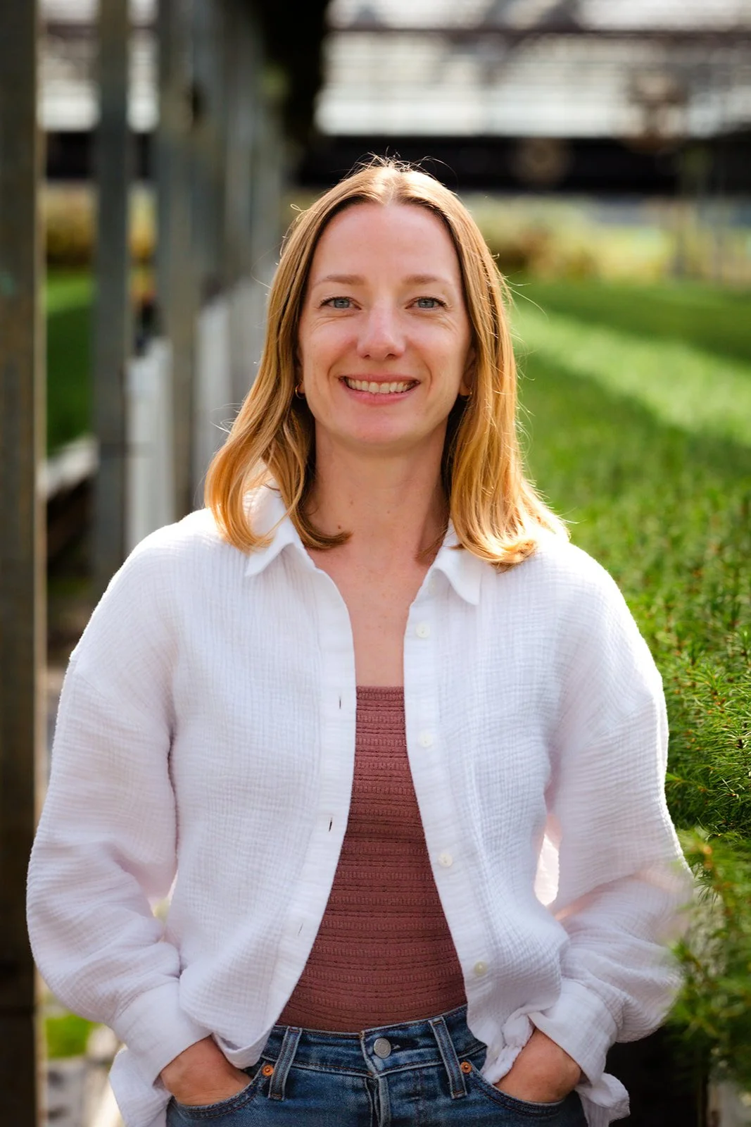 Kelsey Webb with shoulder-length blonde hair, wearing a white button-up shirt over a mauve top, standing outdoors near greenery and a metal structure, smiling at the camera.