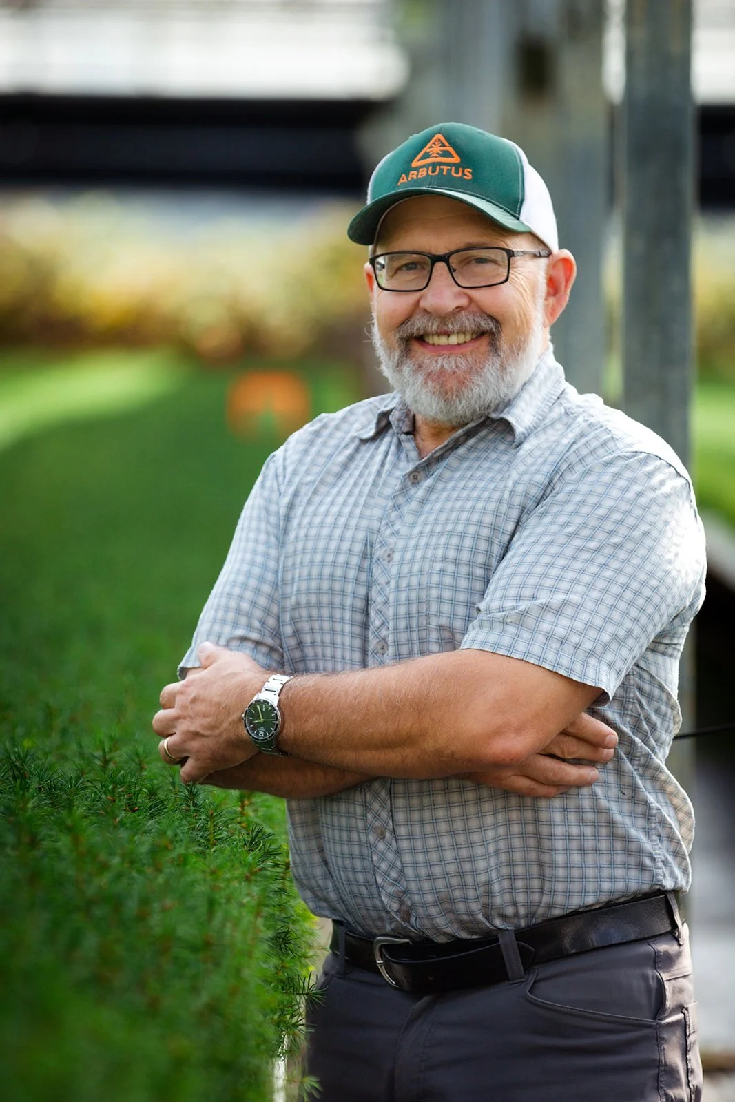 Steven Kiiskila, smiling with glasses and a beard, wearing a green hat and a checkered shirt, standing outdoors with crossed arms behind a green hedge.