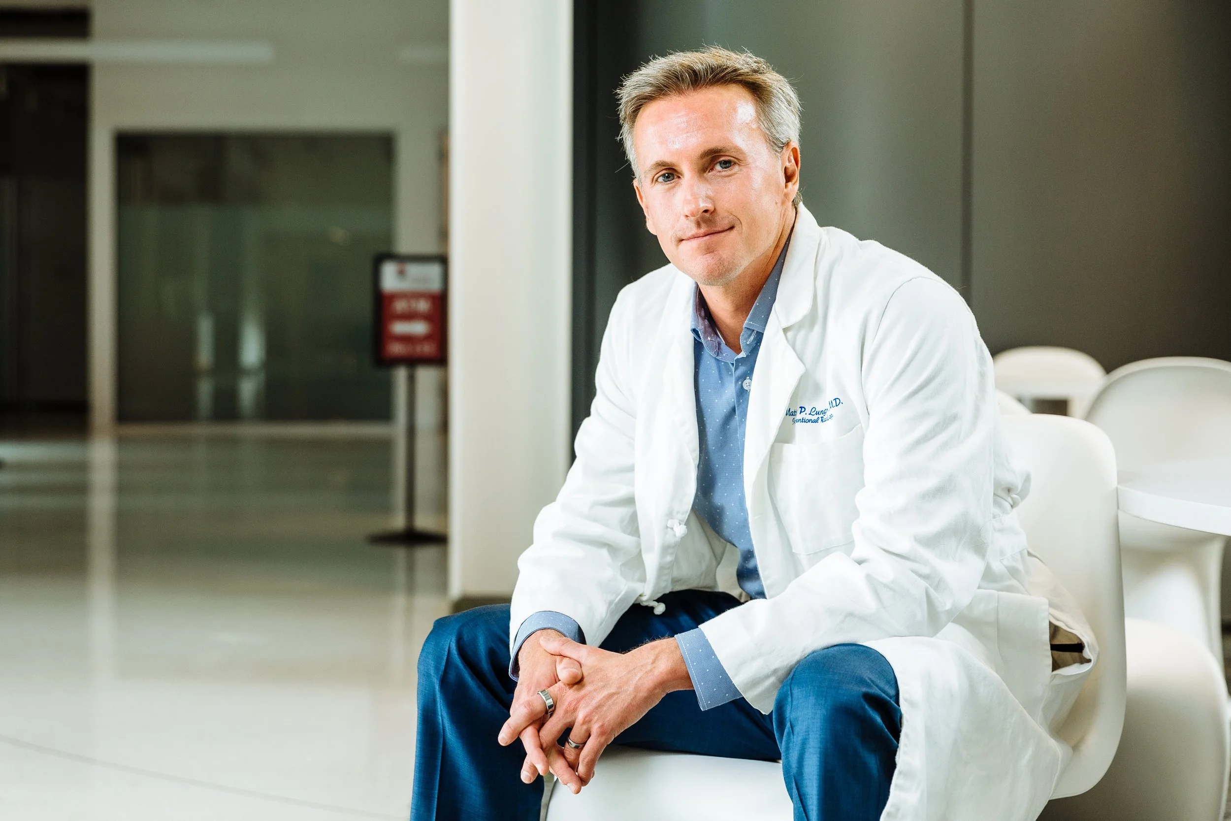 A male doctor in a white coat sitting in a hospital or clinic lobby, with a confident expression.
