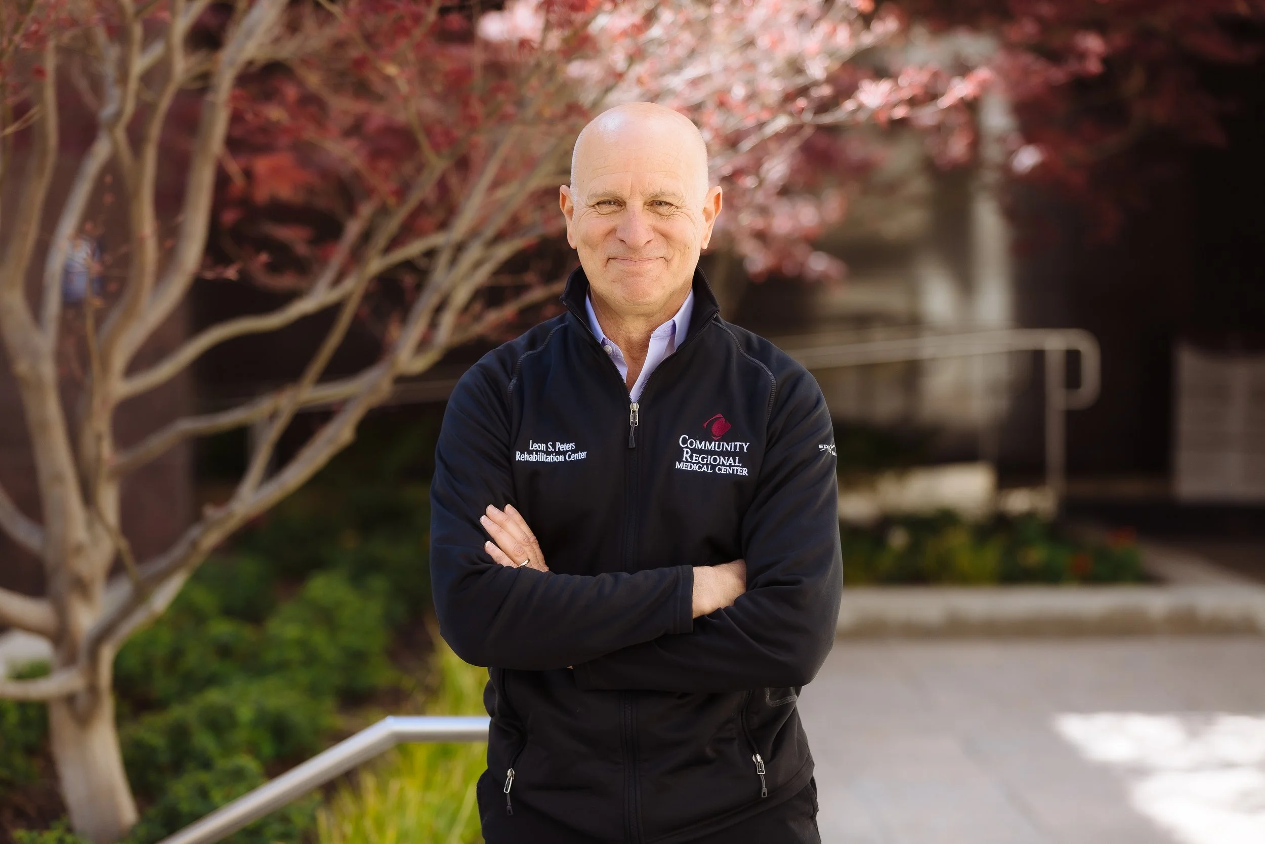 A smiling bald man wearing a black jacket with the logo of a medical center, standing outdoors near a tree with red leaves and a building in the background.