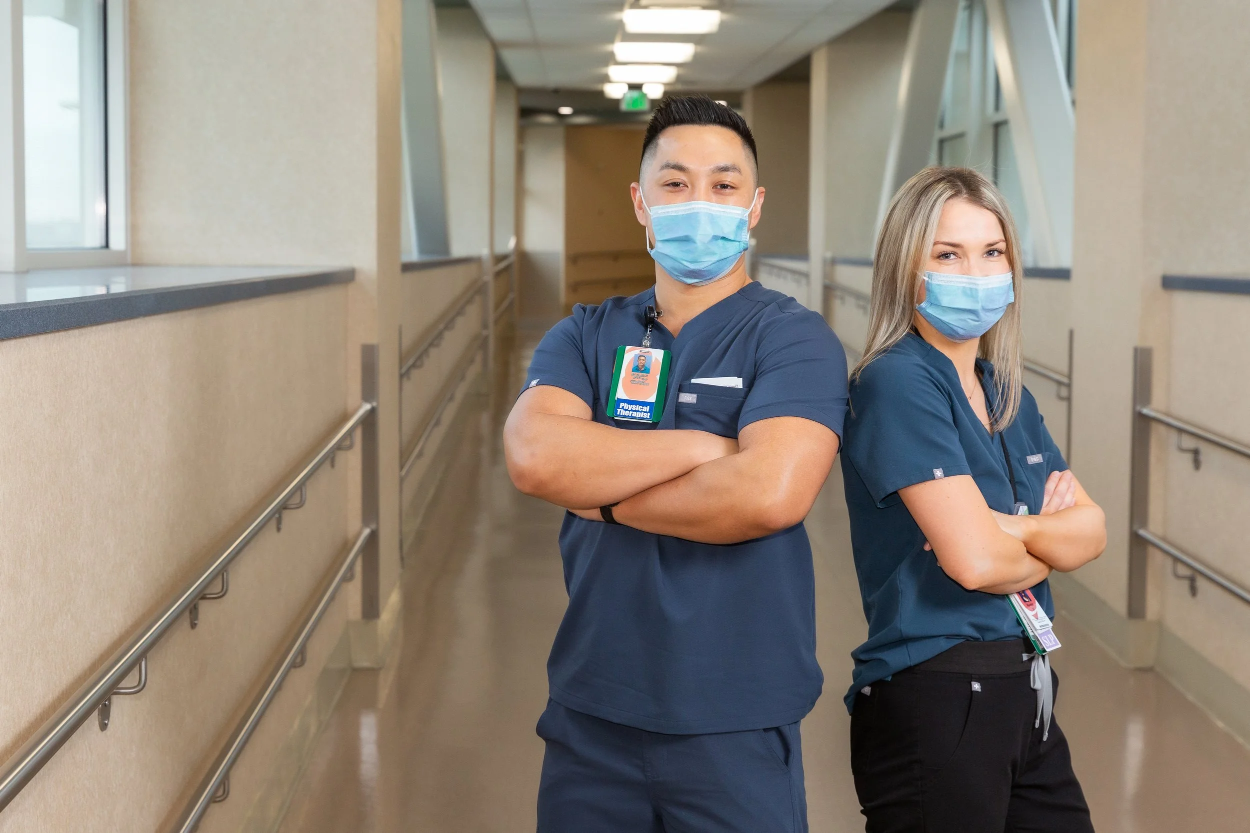 Two healthcare professionals standing back-to-back in a hospital corridor, both wearing blue scrubs and face masks, with arms crossed.