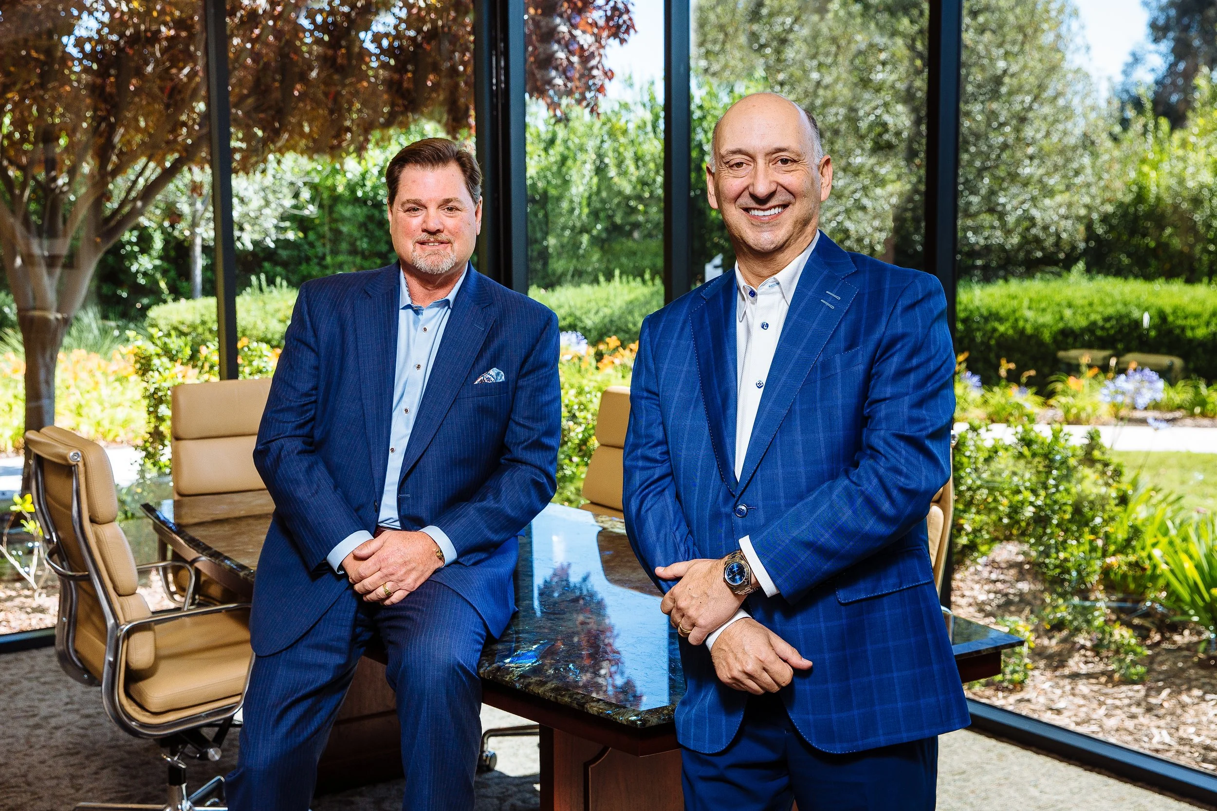 Two men in blue suits standing inside a conference room with large windows revealing an outdoor garden with trees and bushes.
