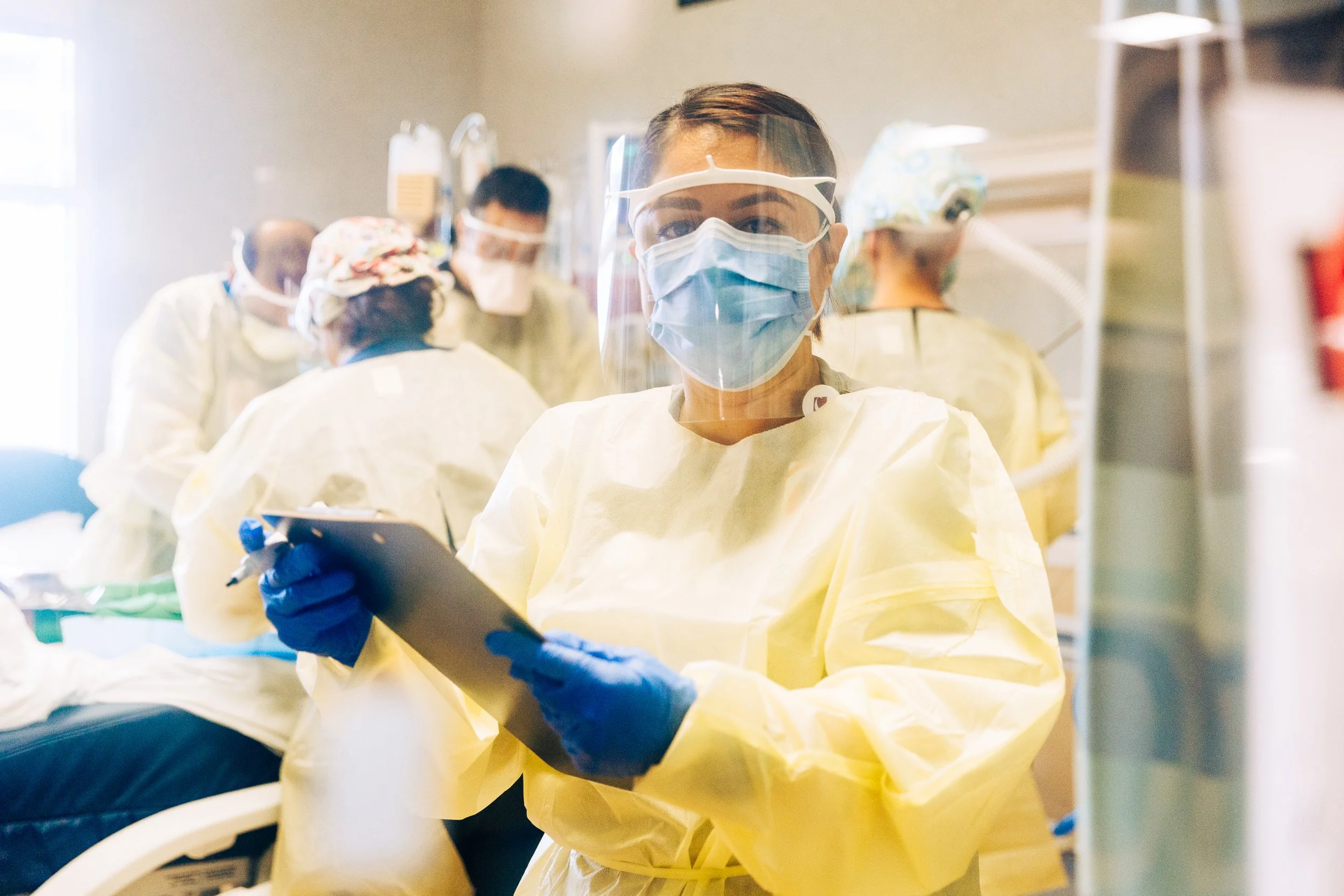 Healthcare workers in protective gear, including masks, gloves, and gowns, inside a medical facility, with one worker holding a tablet.
