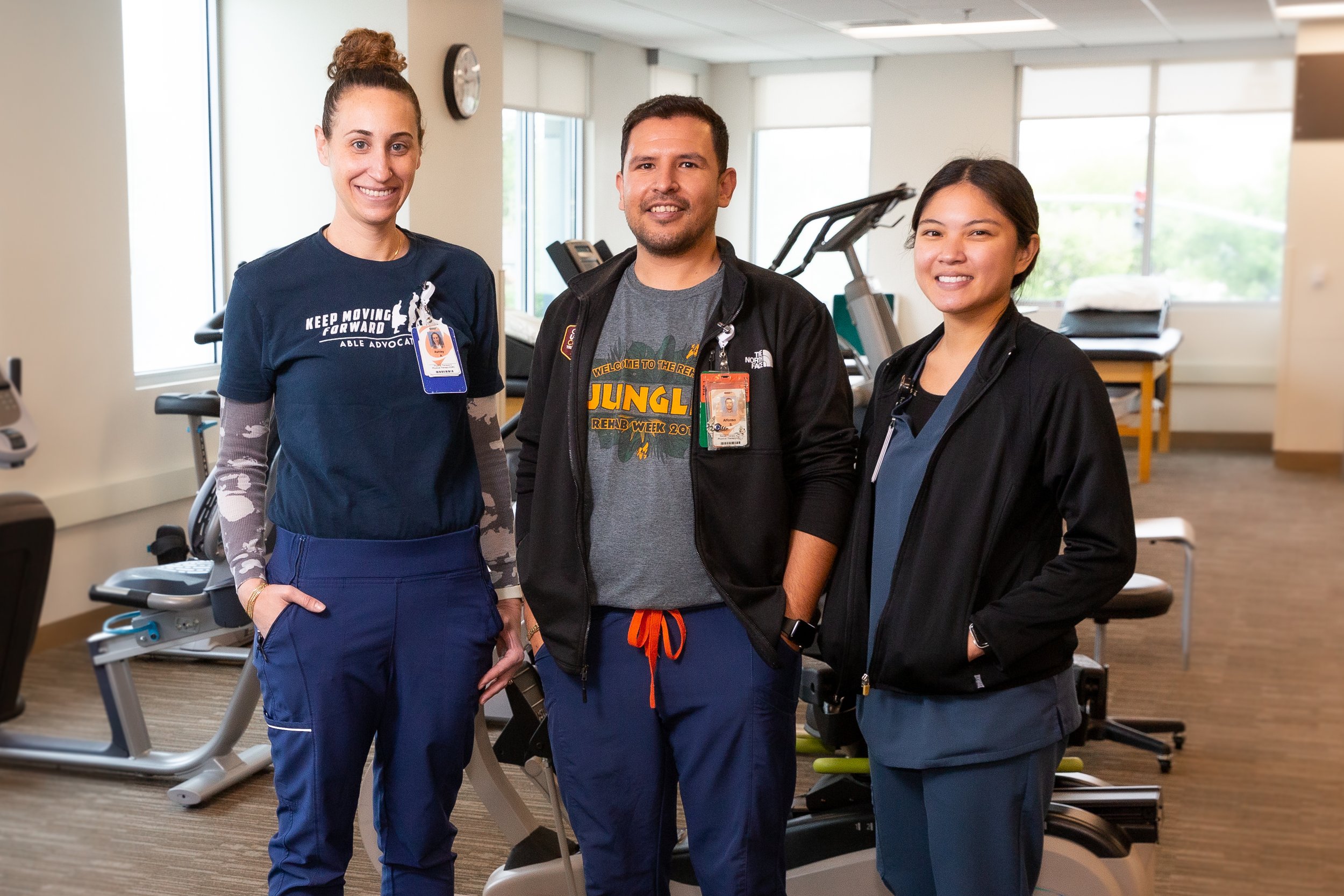 Three healthcare workers standing together in a medical facility gym, smiling, with exercise equipment visible in the background.