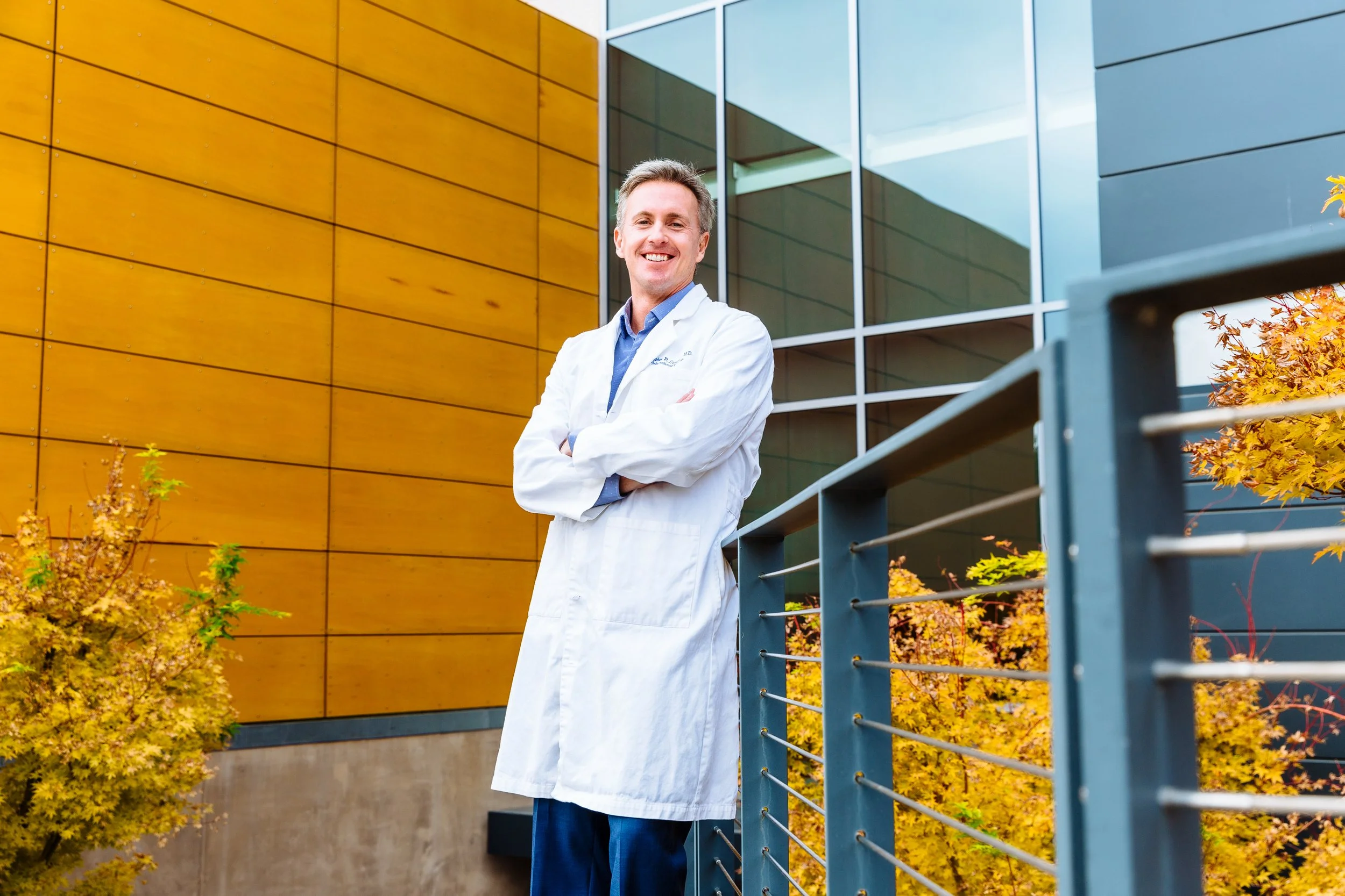 A smiling male doctor in a white coat stands outdoors in front of a modern building with large glass windows and yellow-orange walls, with autumn-colored trees nearby.