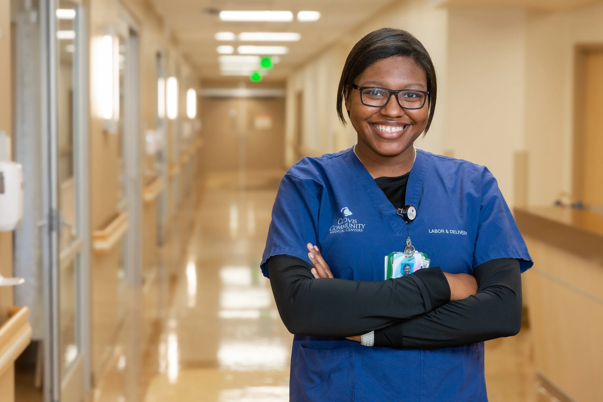 A smiling female healthcare worker in blue scrubs standing with arms crossed inside a hospital corridor.