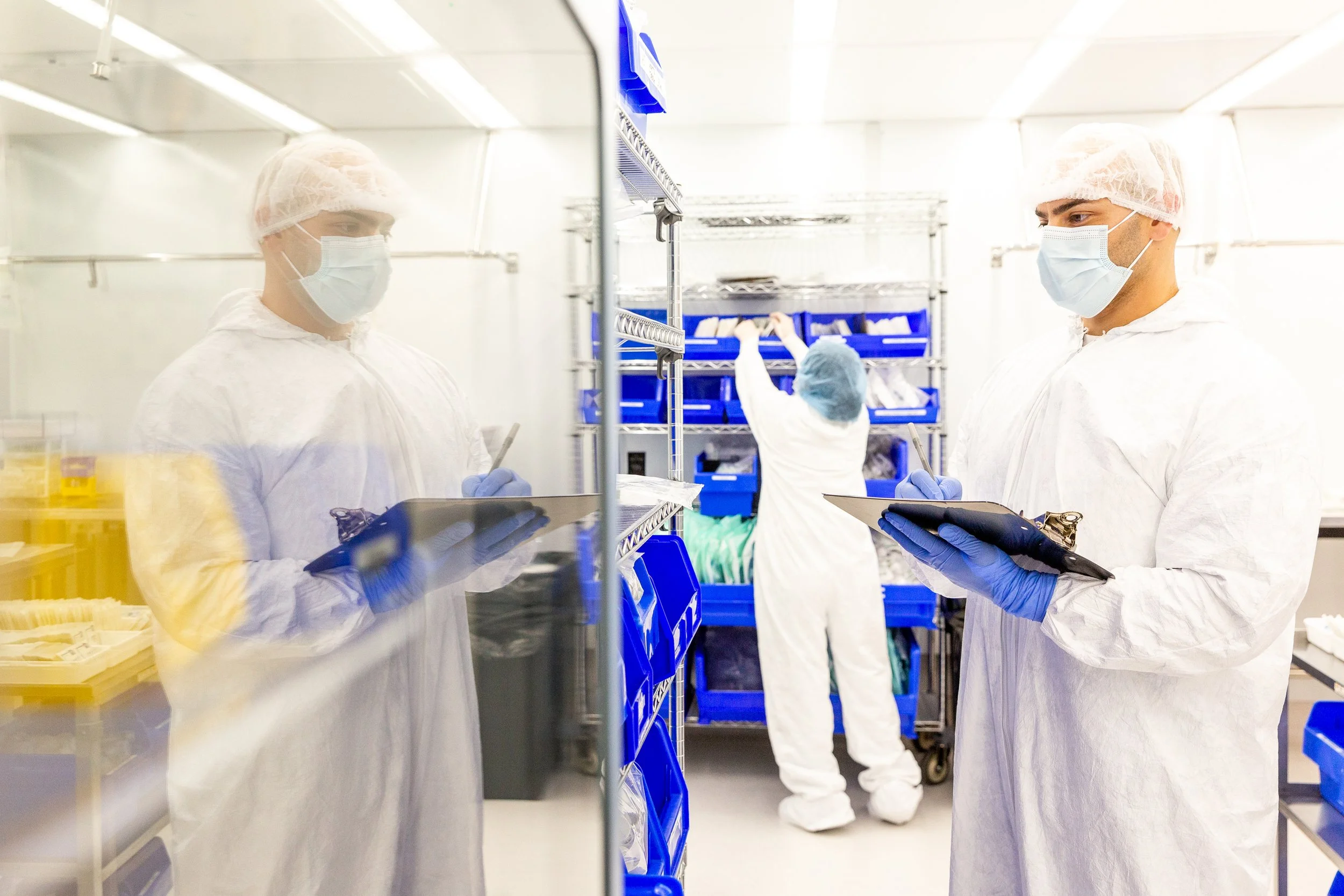 Two scientists in protective suits, face masks, and gloves working in a laboratory. One is taking notes on a clipboard, and the other is working on shelves with blue storage bins behind a glass partition.