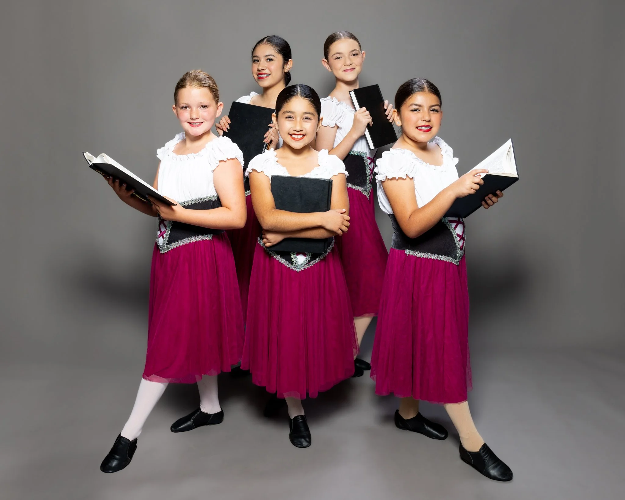 Six young girls dressed in traditional attire holding music scores and books, standing against a gray background.