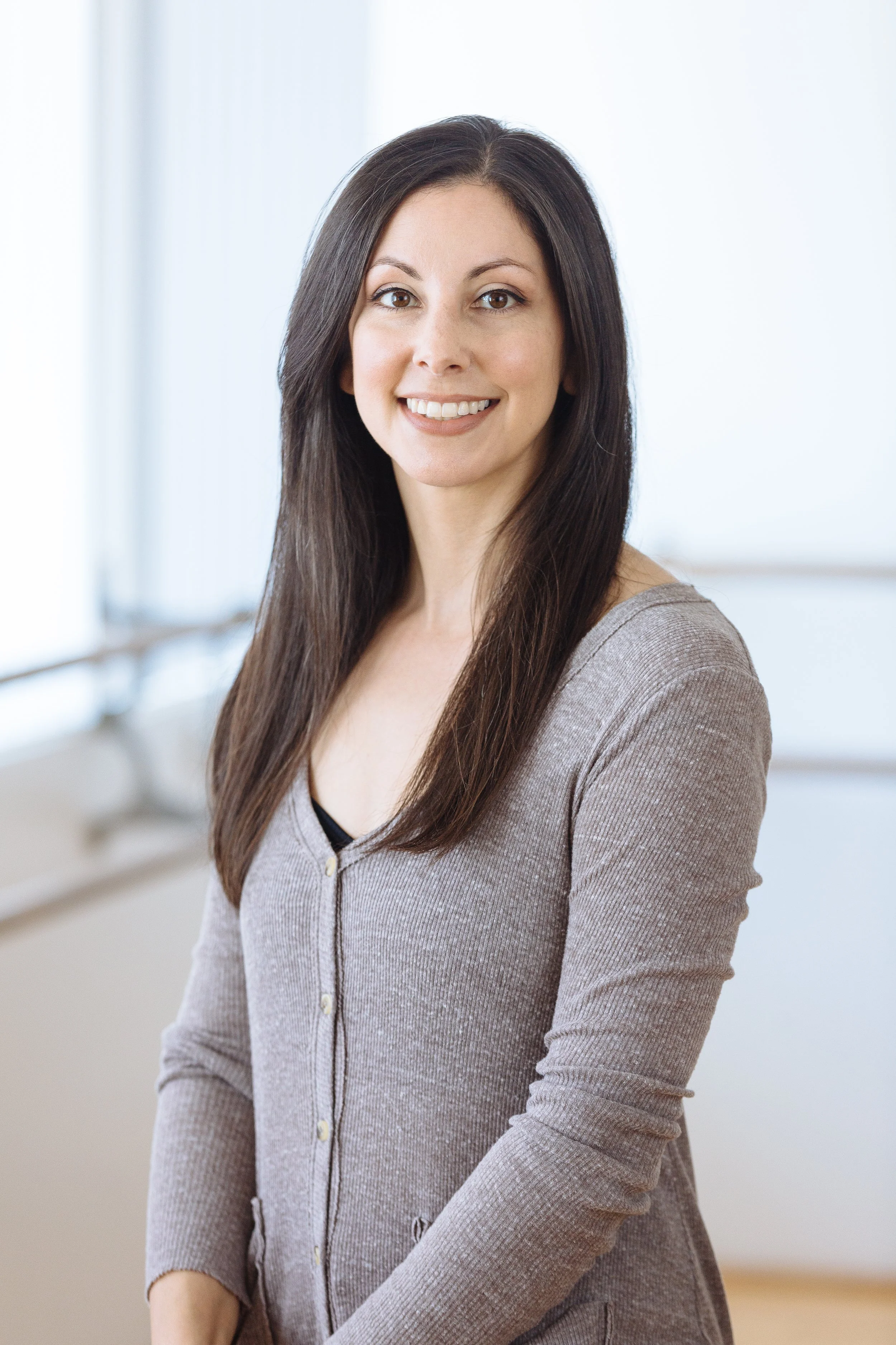 A woman with long dark hair and a beige cardigan smiling at the camera inside a bright room.