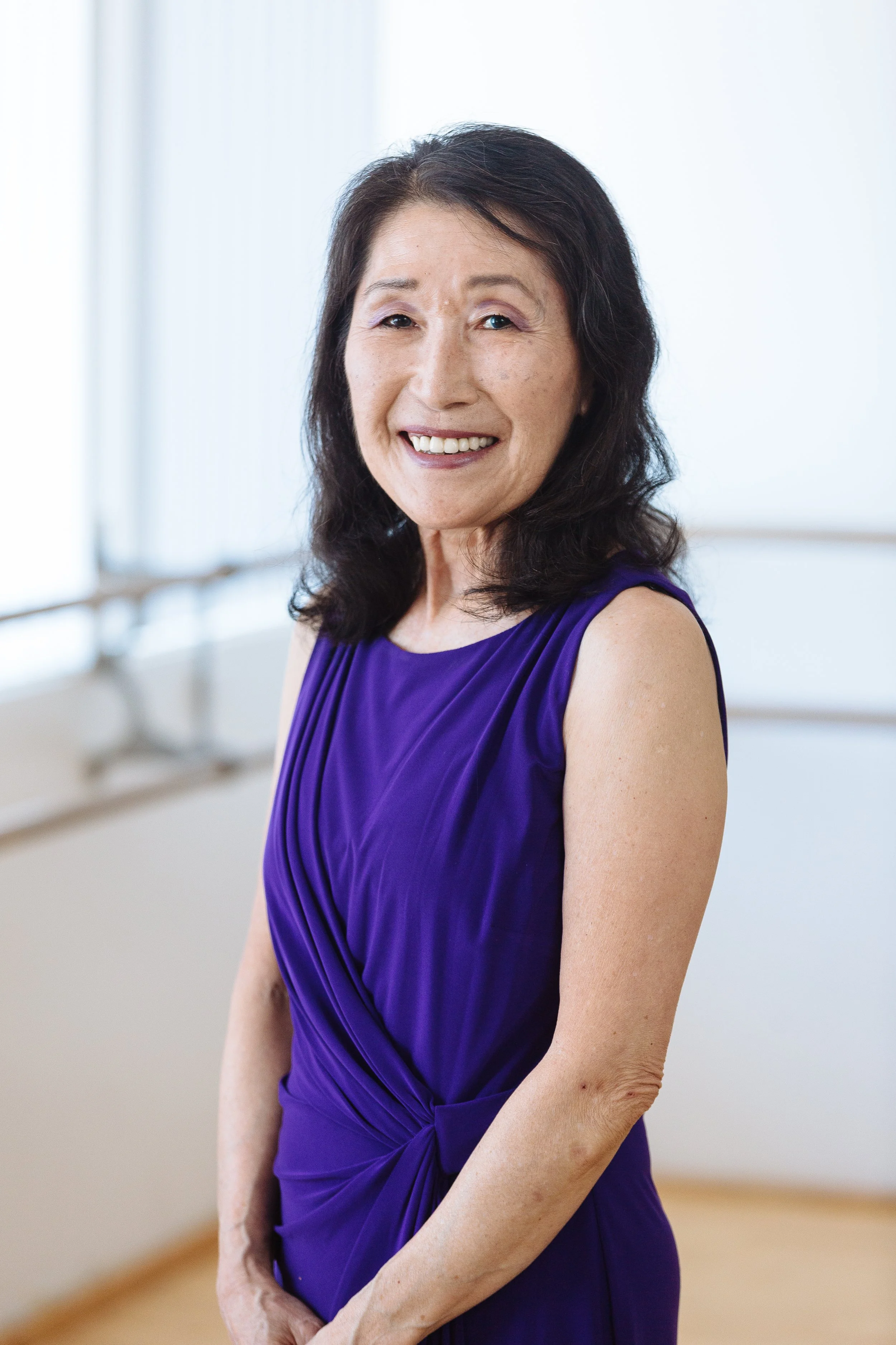 Portrait of a smiling woman in a purple dress standing in a bright room with large windows.
