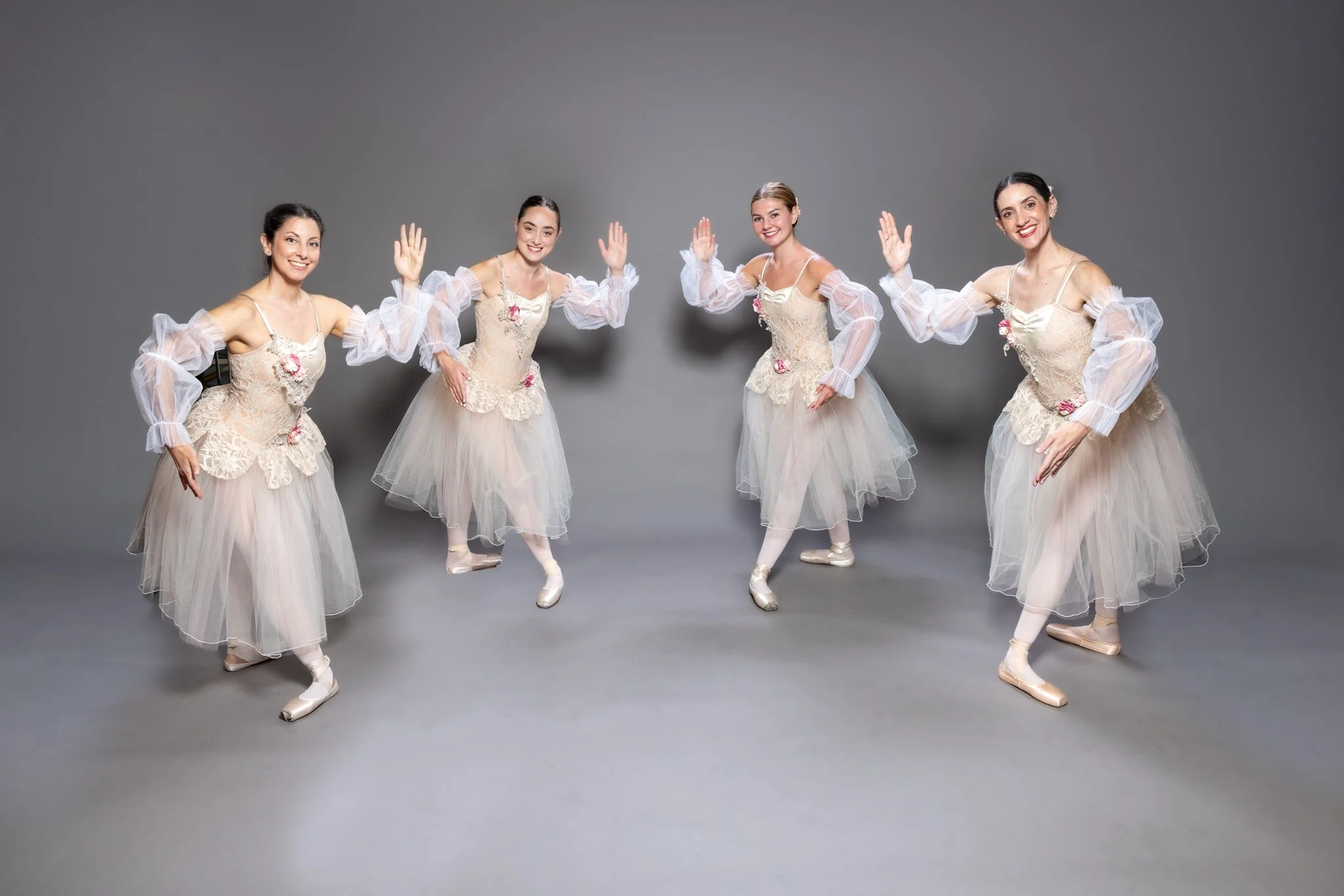 Four women dressed as ballerinas standing in a line, smiling, and waving their hands, against a plain gray background.