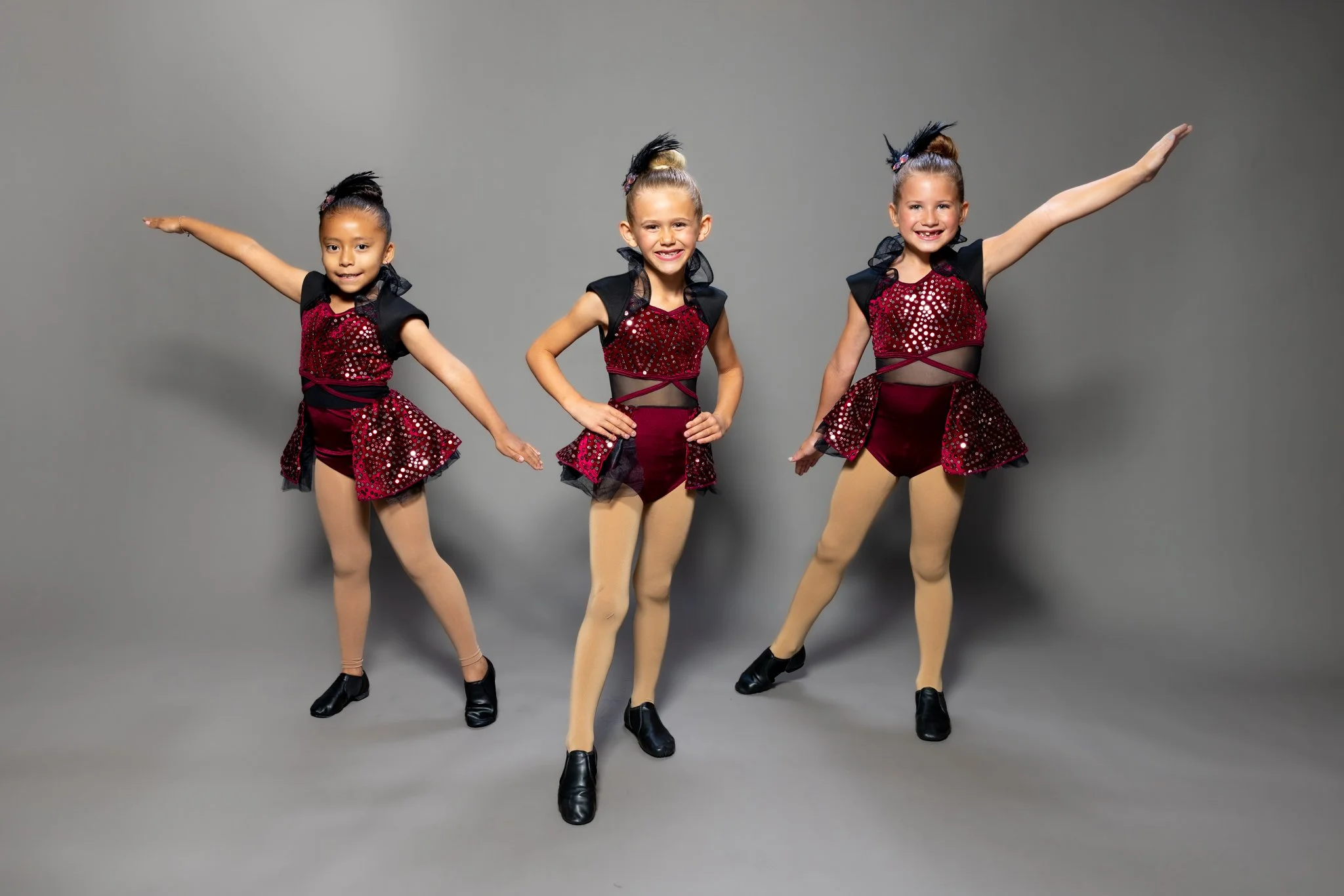 Three young girls in sparkly red and black dance costumes posing against a gray background.