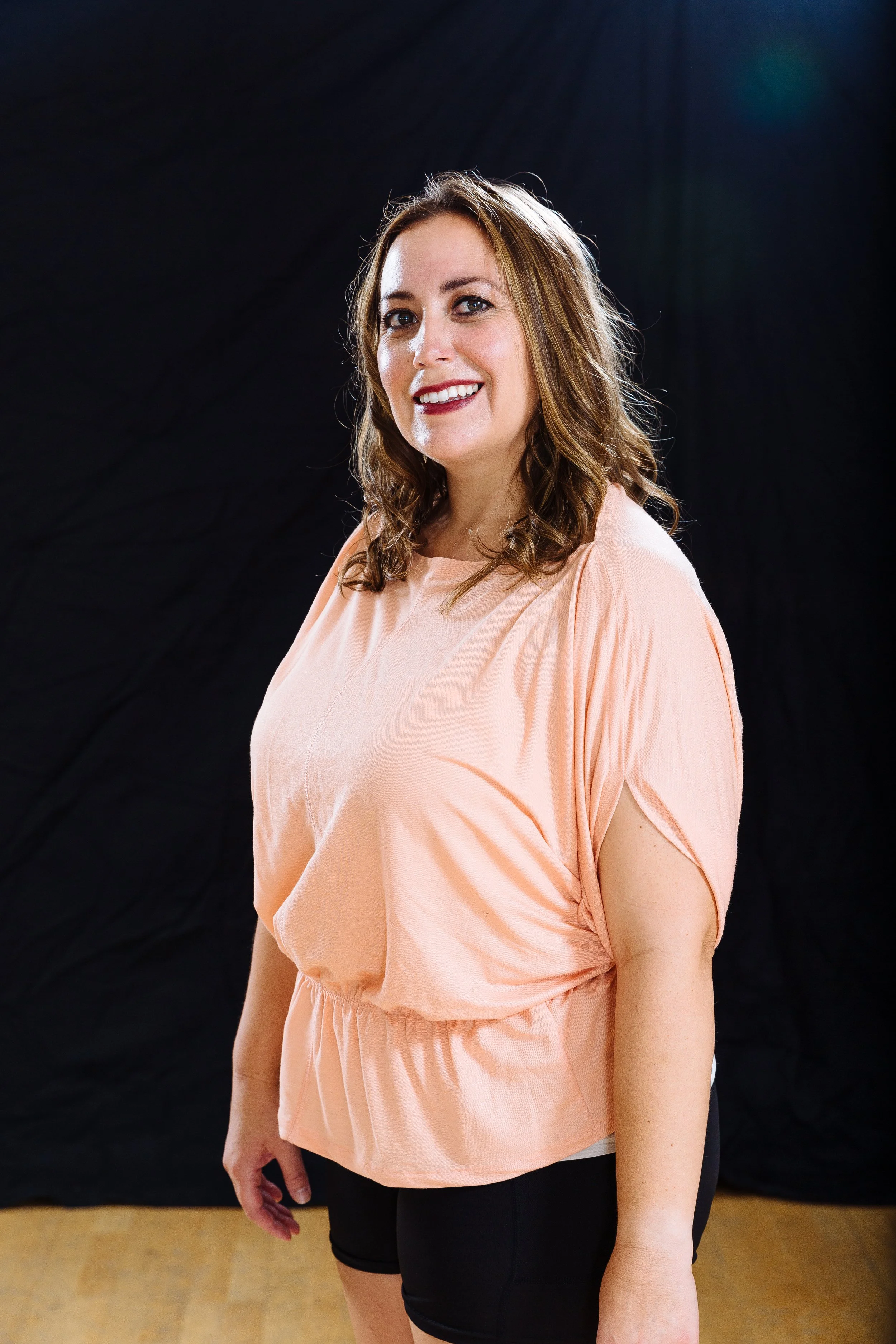 A woman with shoulder-length brown hair, smiling, wearing a peach-colored top and black shorts, standing against a dark background.